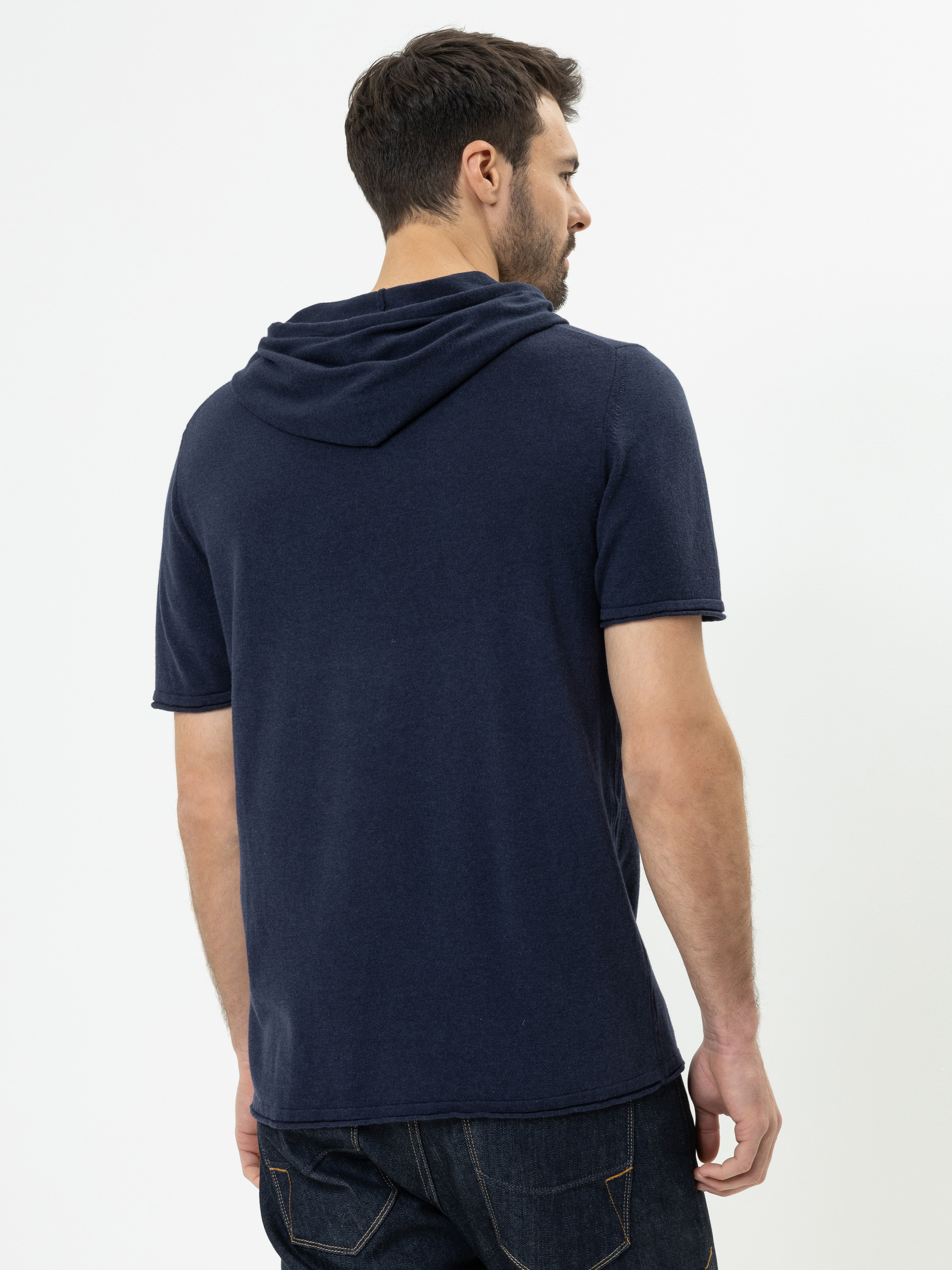 Ein Mann steht mit dem Rücken zur Kamera, trägt ein marineblaues, kurzärmeliges Kapuzenshirt und dunkle Jeans vor einem schlichten weißen Hintergrund.