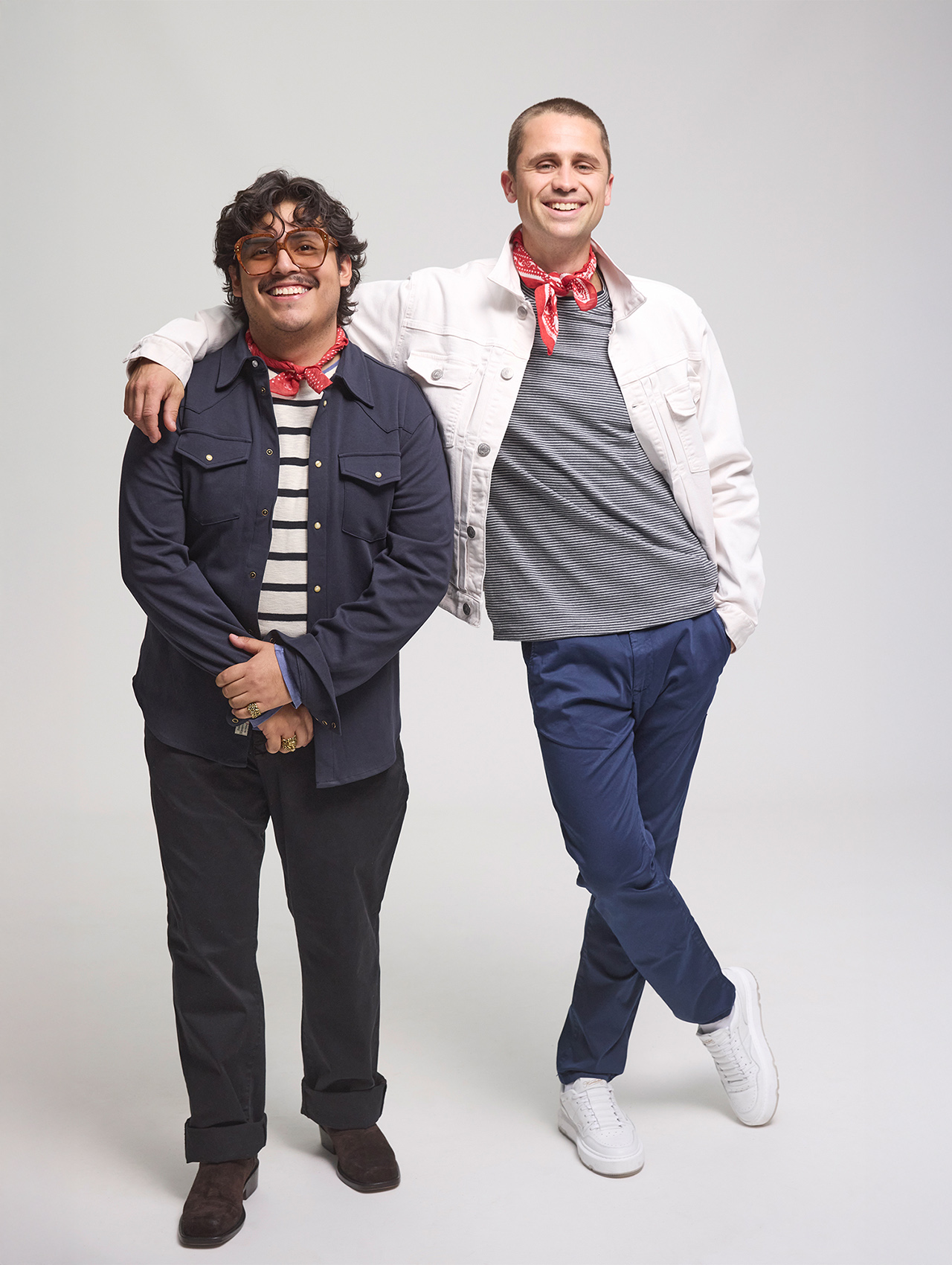 Zwei Männer stehen lächelnd in einem Studio mit einfarbigem Hintergrund. Der eine trägt eine Brille, eine marineblaue Jacke, ein gestreiftes Hemd und eine dunkle Hose. Der andere trägt eine weiße Jacke, ein graues Hemd, eine blaue Hose und weiße Tur