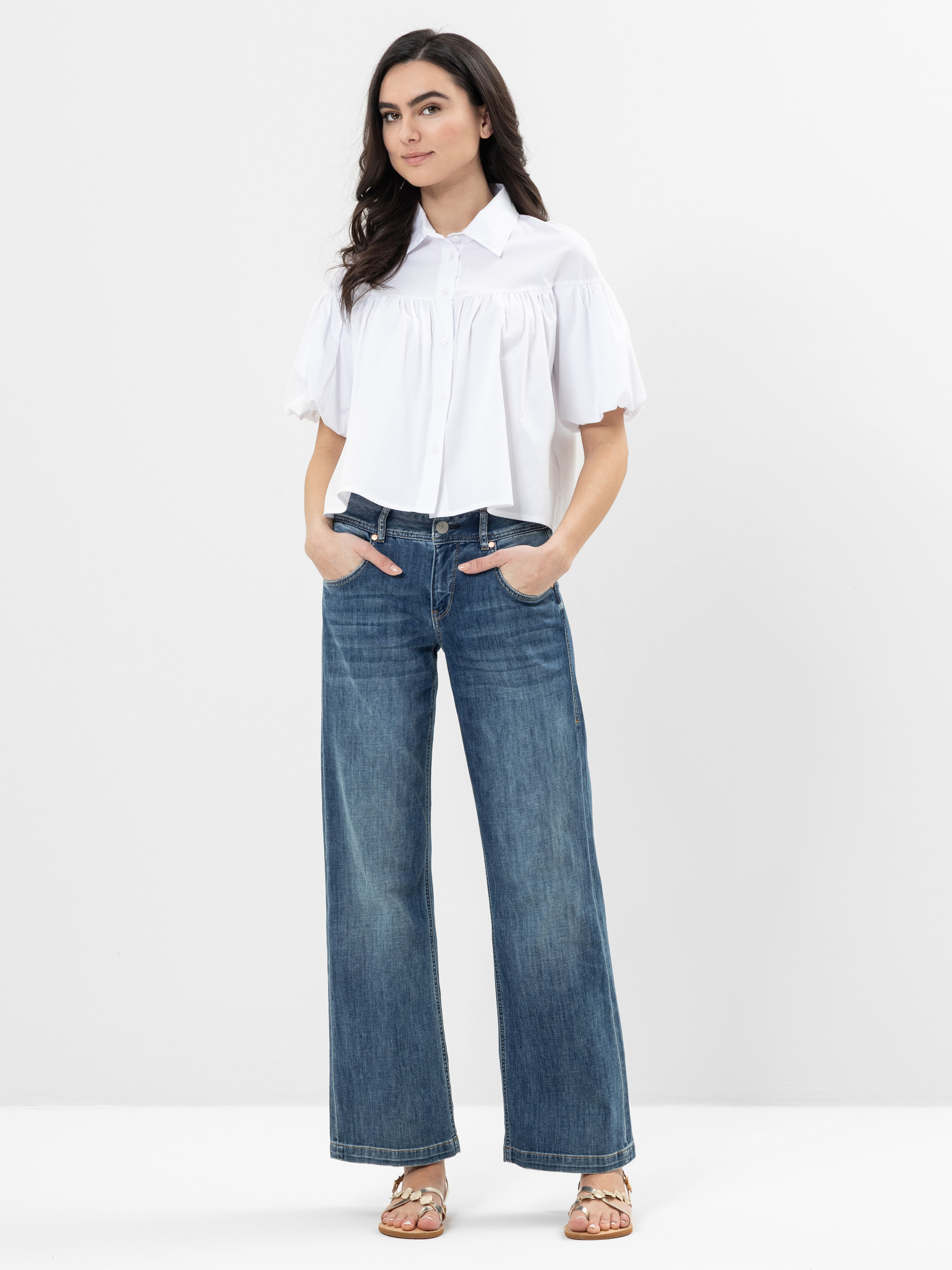 Eine Frau steht vor einem schlichten weißen Hintergrund und trägt eine weite weiße Bluse mit kurzen Puffärmeln, eine weit geschnittene Bluejeans und Sandalen. Sie hat langes dunkles Haar und steckt die Hände in die Taschen ihrer Jeans.