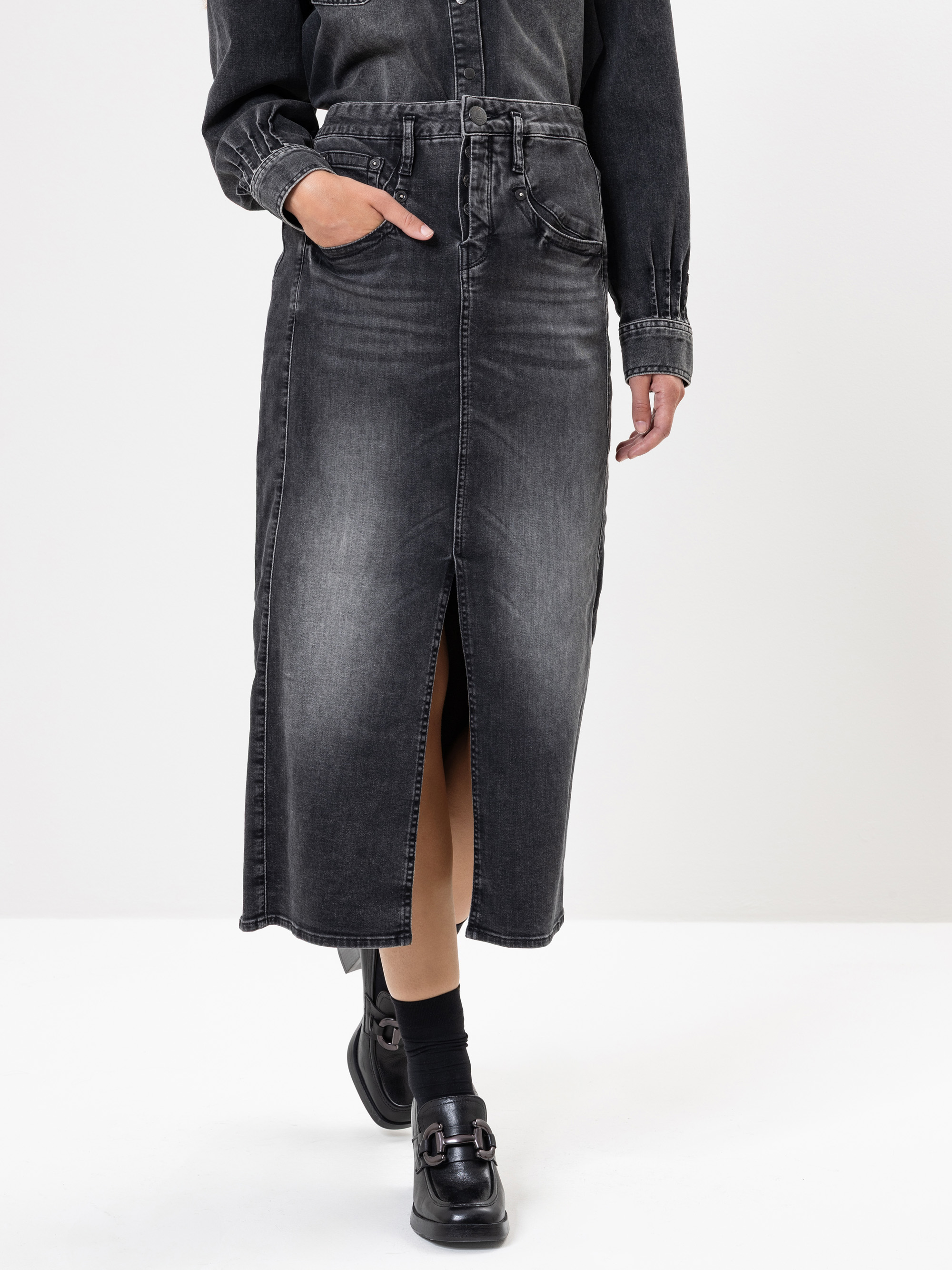 Eine Person trägt einen hochtaillierten schwarzen Midirock aus verwaschenem Jeansstoff mit einem Schlitz an der Vorderseite, dazu ein passendes Jeanshemd, schwarze Socken und schwarze Schnallenschuhe. Die Person hat eine Hand in ihrer Tasche.