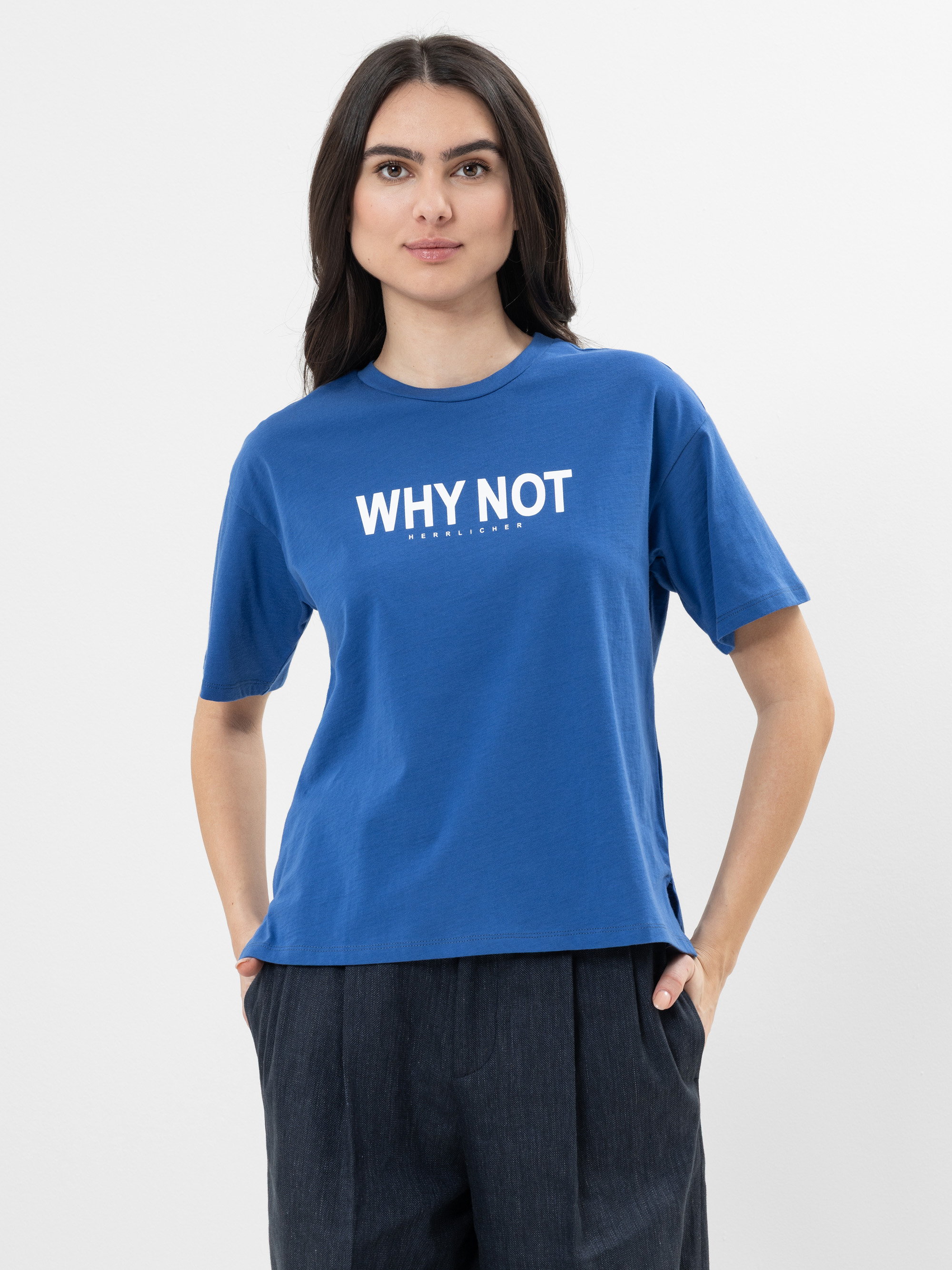 Eine Frau mit langen dunklen Haaren steht vor einem weißen Hintergrund, trägt ein blaues T-Shirt mit dem Text WHY NOT in weißer Schrift auf der Vorderseite und eine dunkle Hose. Sie blickt nach vorne und hat die Hände in den Taschen.