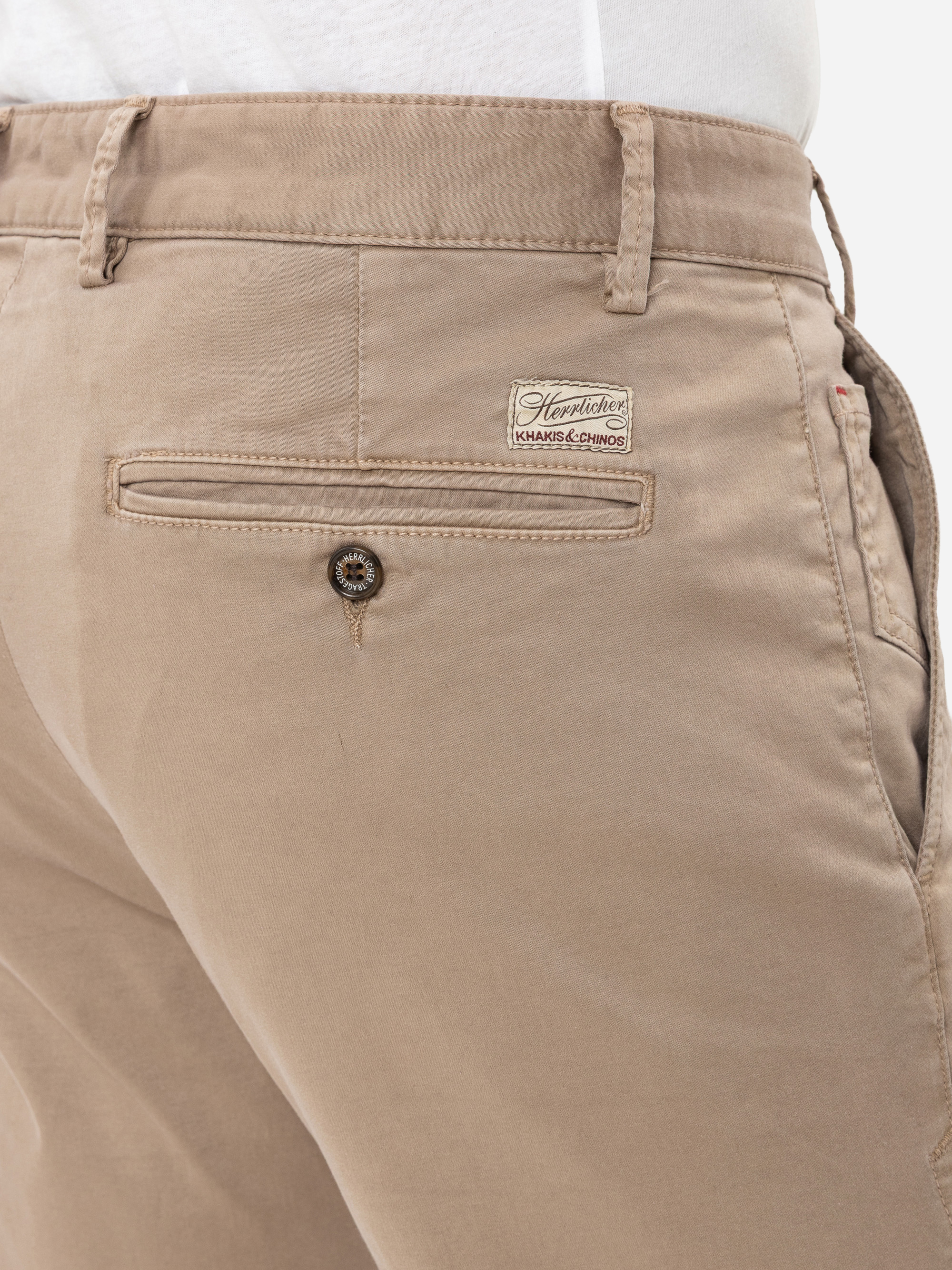 Nahaufnahme des Rückens einer Person, die eine beigefarbene Chinohose mit einer geknöpften Gesäßtasche, Gürtelschlaufen und einem weißen Etikett mit der Aufschrift "Handpicked HAND-CHINOS" über der Tasche trägt.