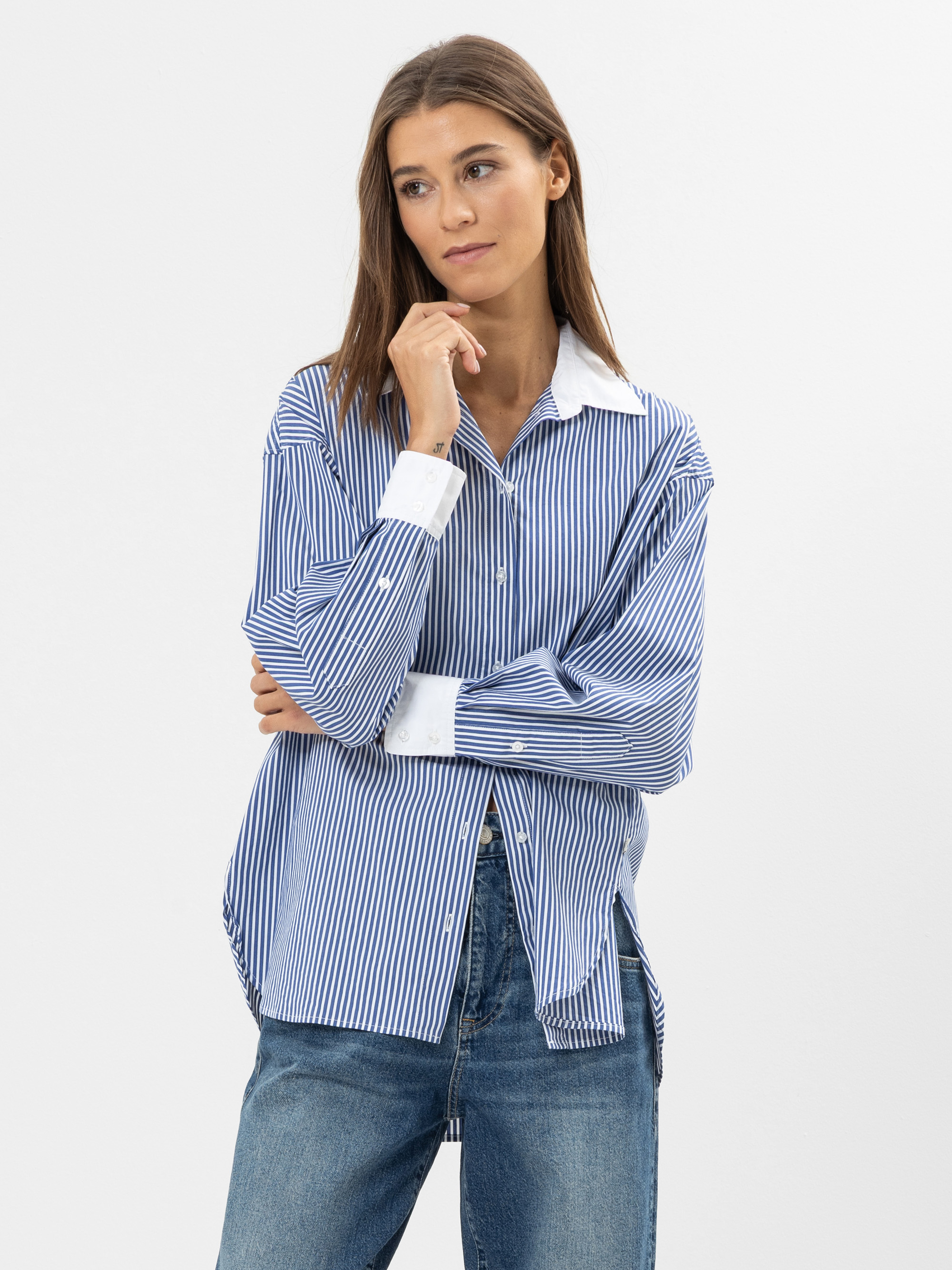 Eine Frau mit langen braunen Haaren trägt ein lockeres blau-weiß gestreiftes Button-up-Hemd und blaue Jeans. Sie steht mit nachdenklichem Gesichtsausdruck vor einem schlichten weißen Hintergrund und berührt mit der Hand ihr Kinn.