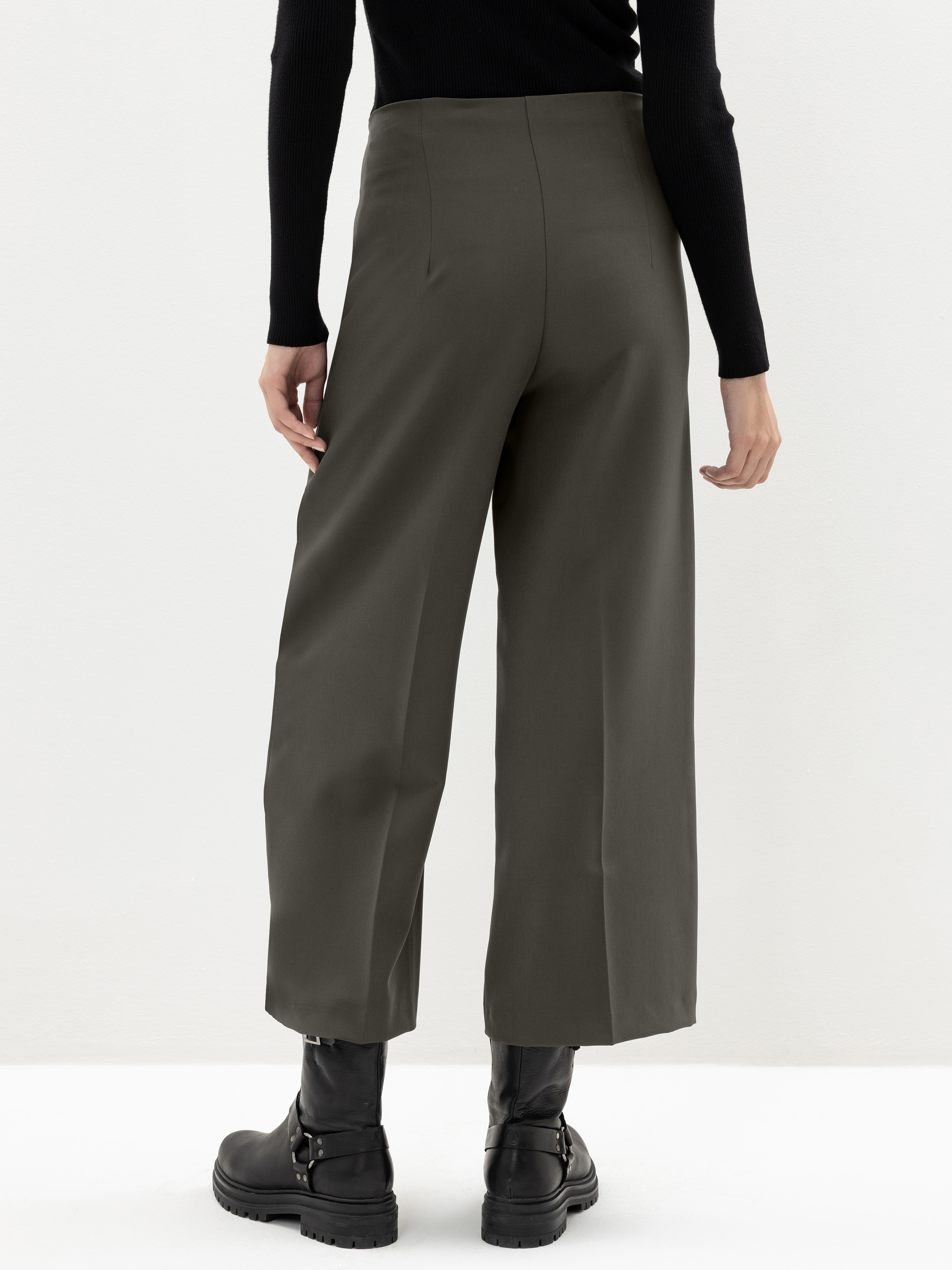 Eine Person steht vor einem hellen Hintergrund und trägt eine olivgrüne Hose mit weitem Bein, ein schwarzes langärmeliges Oberteil und schwarze Stiefeletten. Das Foto ist von hinten aufgenommen und zeigt die Passform und Länge der Hose.