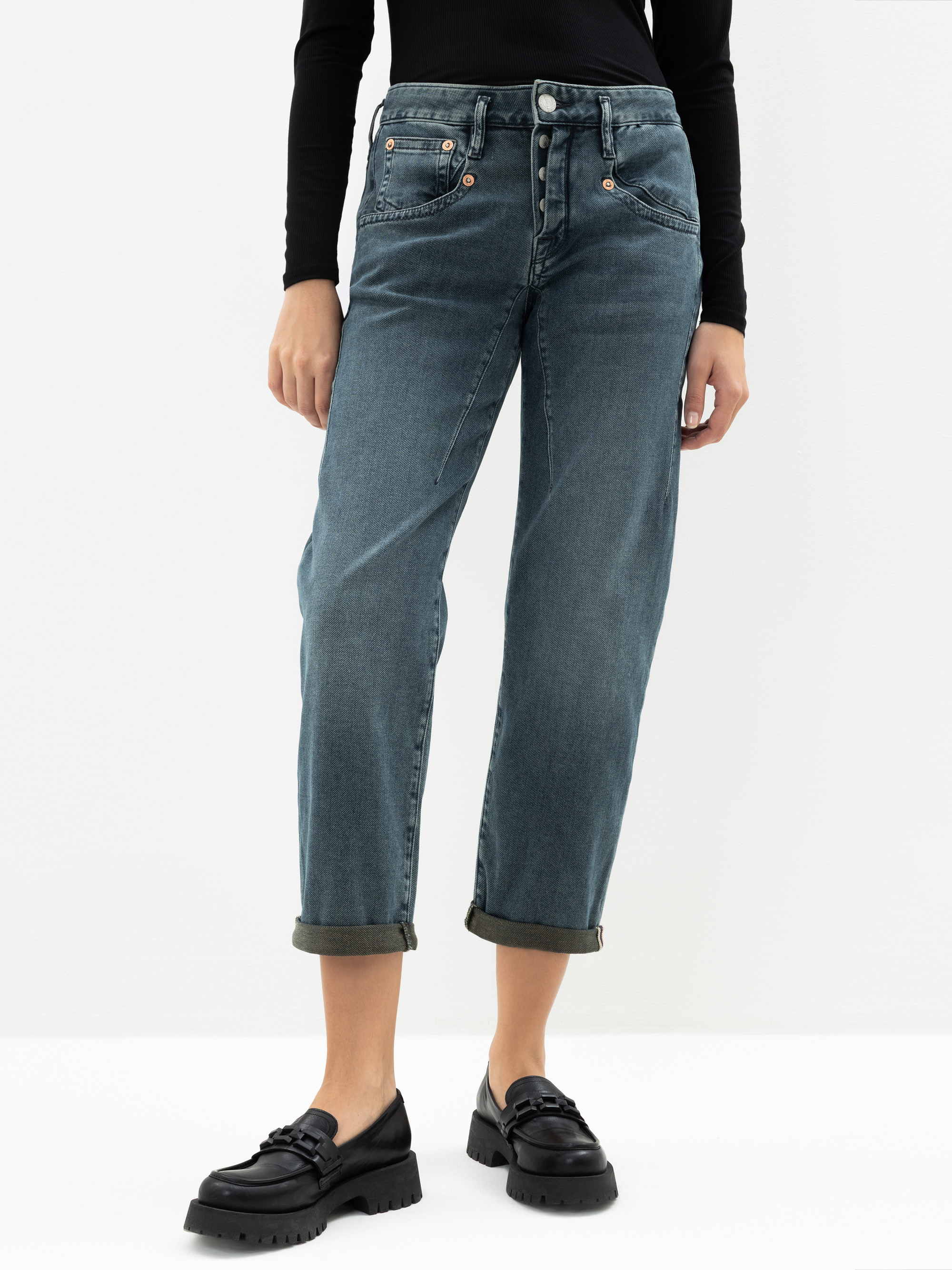 Eine Person, die ein schwarzes langärmeliges Oberteil, blaue Jeans mit Bündchen und schwarze Halbschuhe trägt, steht vor einem schlichten weißen Hintergrund. Das Bild ist von den Schultern abwärts beschnitten.
