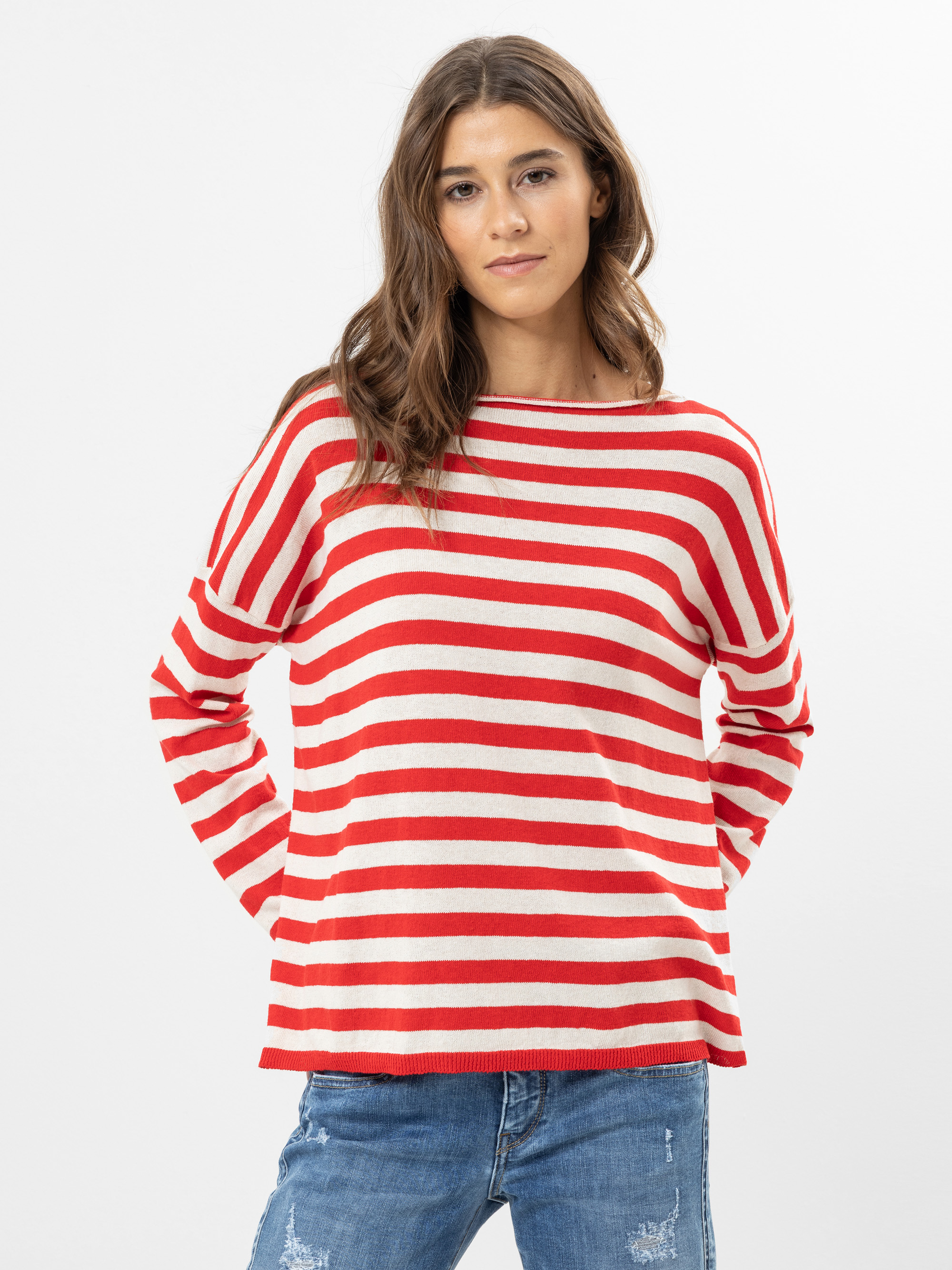Eine Frau mit langen braunen Haaren trägt einen rot-weiß gestreiften Pullover und blaue Jeans und steht mit den Händen in den Taschen vor einem schlichten weißen Hintergrund.