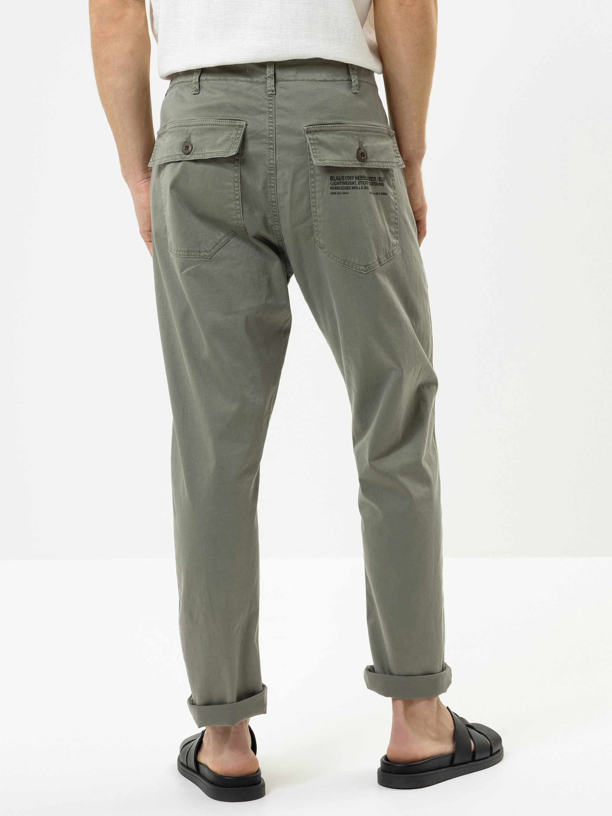 Eine Person trägt eine helle olivgrüne Hose mit aufgerollten Manschetten, schwarze Sandalen und ein weißes Hemd. Das Foto ist von hinten aufgenommen und zeigt zwei geknöpfte Gesäßtaschen, wobei über der rechten Tasche ein Text aufgedruckt ist.