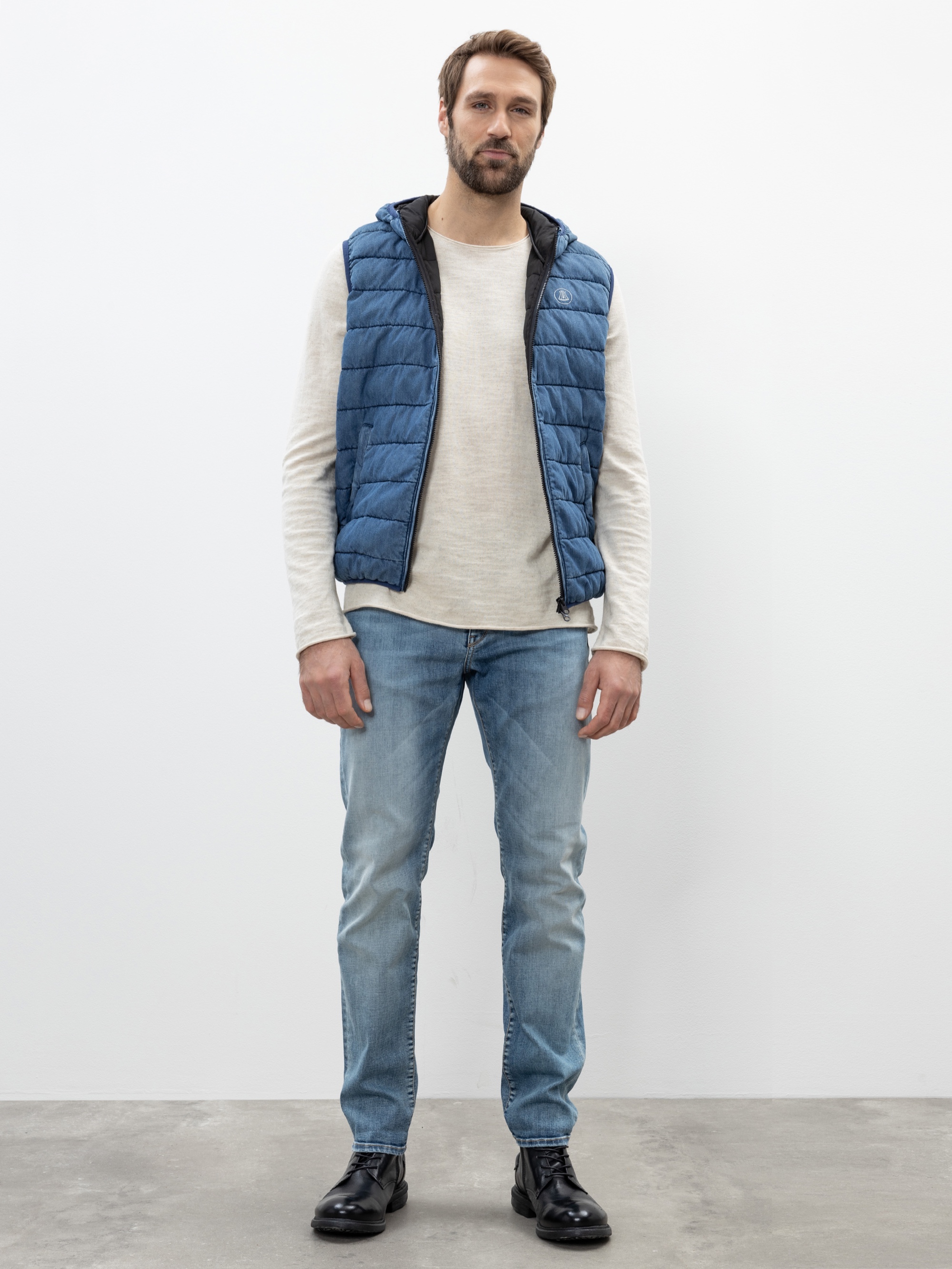 Ein Mann steht vor einem schlichten weißen Hintergrund und trägt eine blaue Pufferweste über einem hellen langärmeligen Hemd, blaue Jeans und schwarze Stiefel. Er hat kurzes Haar und einen Bart, einen entspannten Gesichtsausdruck und die Hände an den