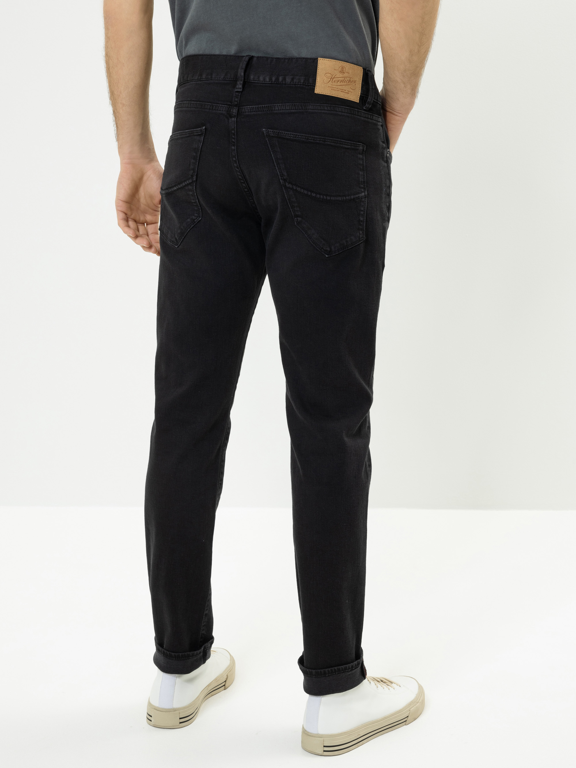 Ein Mann steht mit dem Gesicht nach hinten, trägt eine schwarze Jeans mit Gesäßtaschen und einem hellbraunen Lederaufnäher am Bund, ein dunkles Hemd und weiße Turnschuhe mit hellen Sohlen vor einem schlichten weißen Hintergrund.