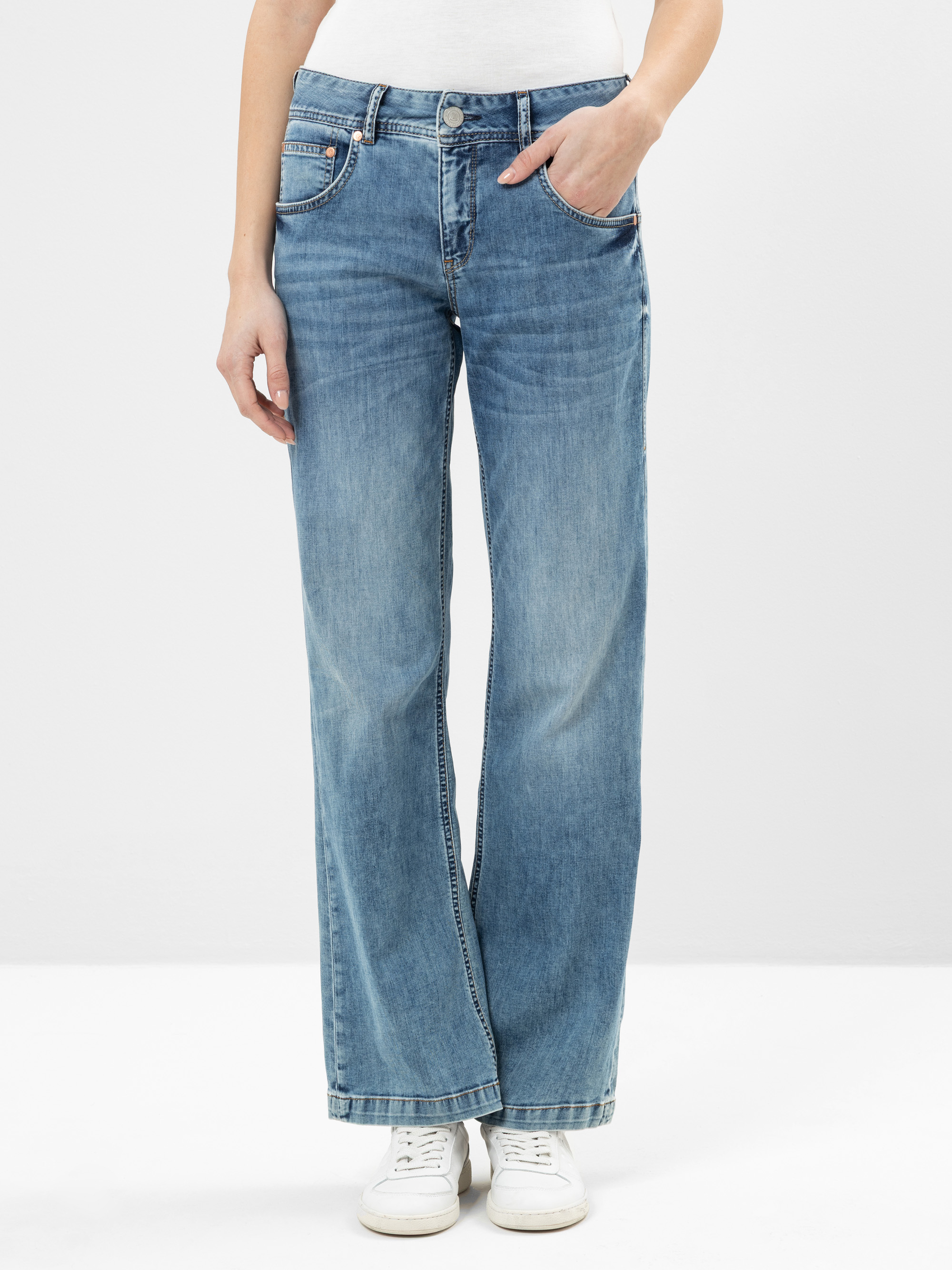 Eine Person in hellblauen Jeans mit weitem Bein, weißem Oberteil und weißen Turnschuhen steht vor einem schlichten weißen Hintergrund und hat eine Hand in der Tasche.