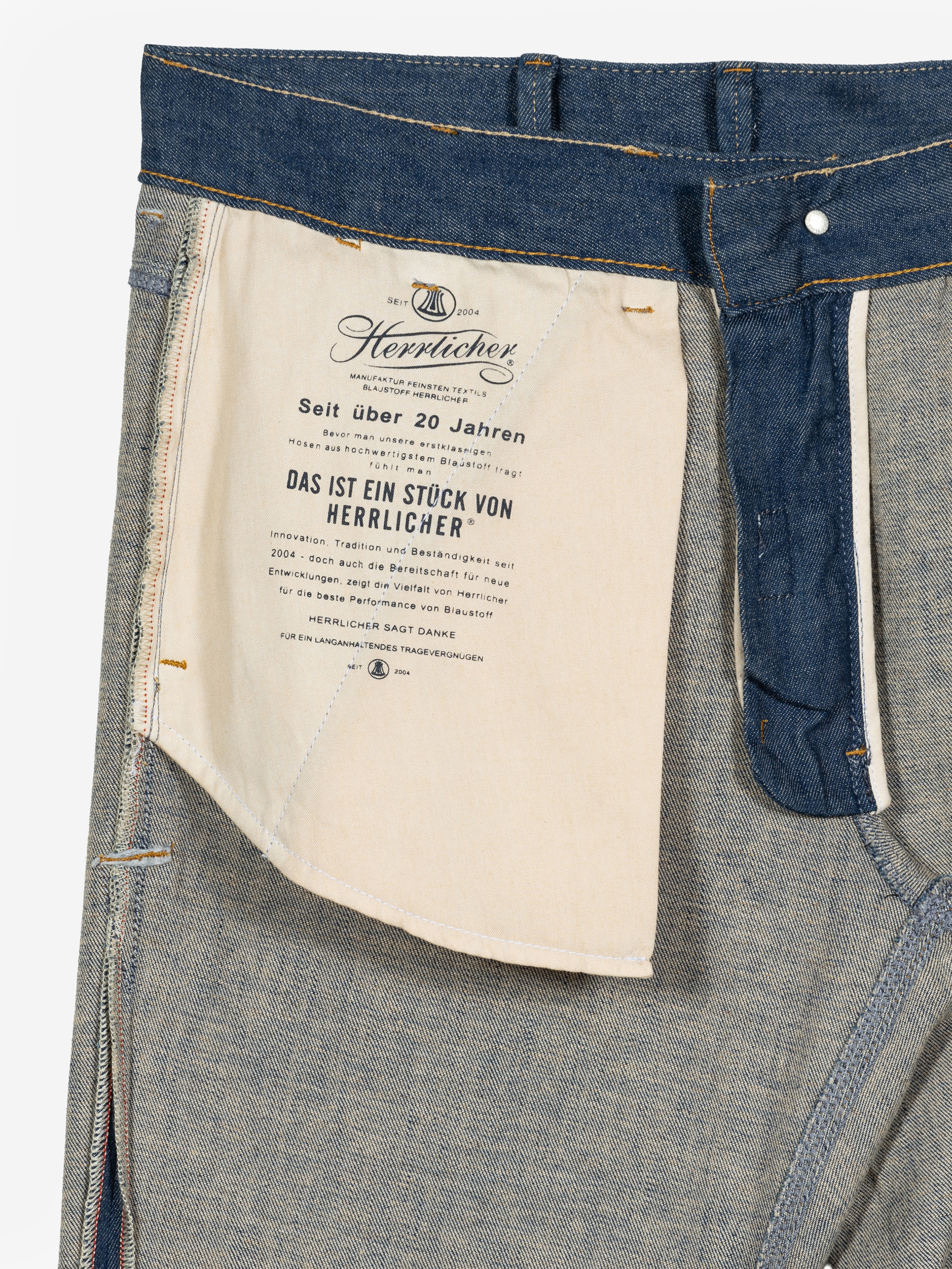 Nahaufnahme der Innenseite einer Jeans, die ein cremefarbenes Taschenfutter mit schwarzem, auf Deutsch gedrucktem Text, Jeansstoff, Nähte sowie einen Teil des Bunds und des Hosenschlitzes zeigt.