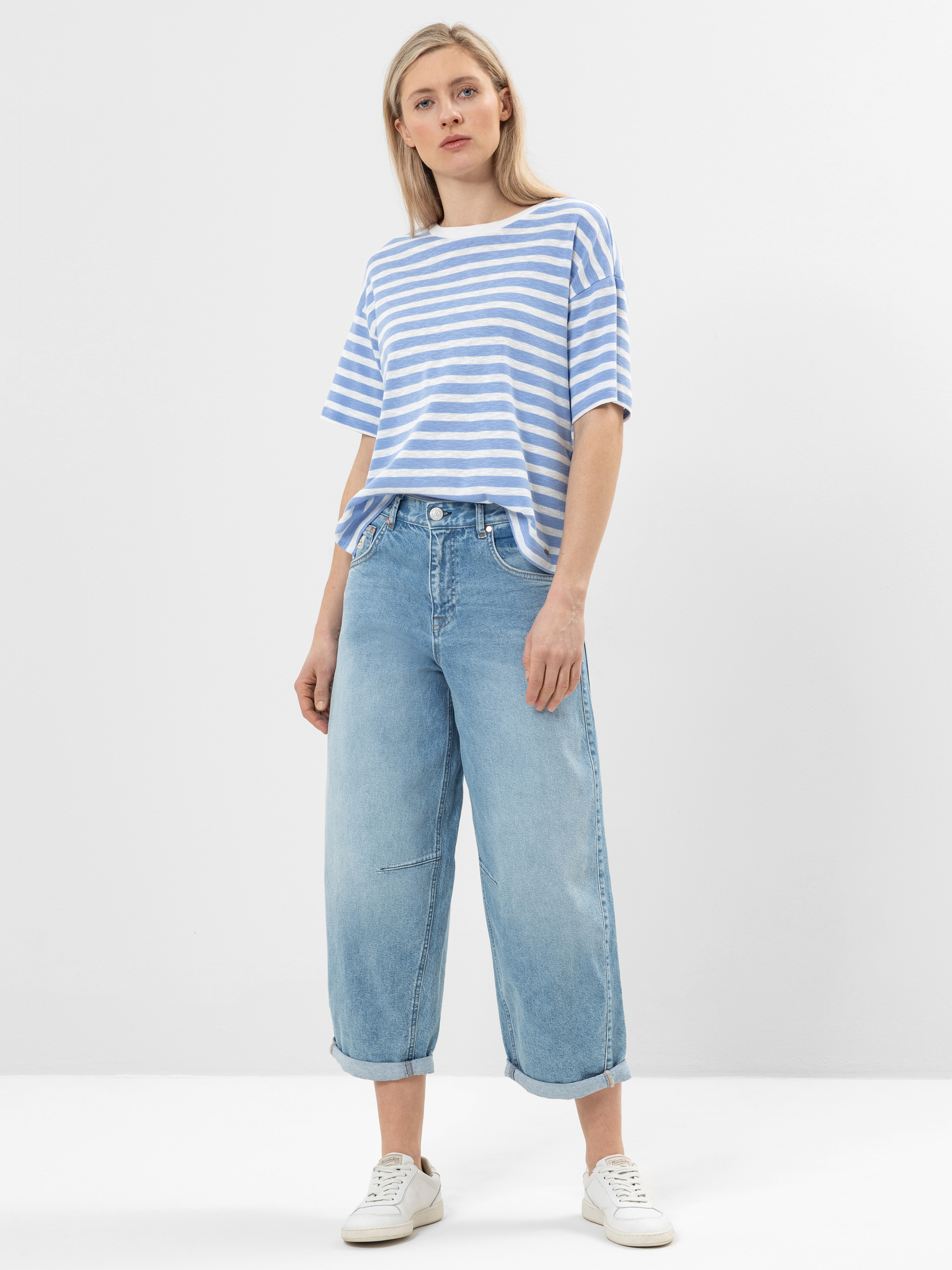 Eine Frau steht vor einem schlichten weißen Hintergrund und trägt ein blau-weiß gestreiftes T-Shirt, eine locker sitzende hellblaue Jeans mit aufgerollten Bündchen und weiße Turnschuhe. Sie hat glattes blondes Haar und einen neutralen Gesichtsausdruc