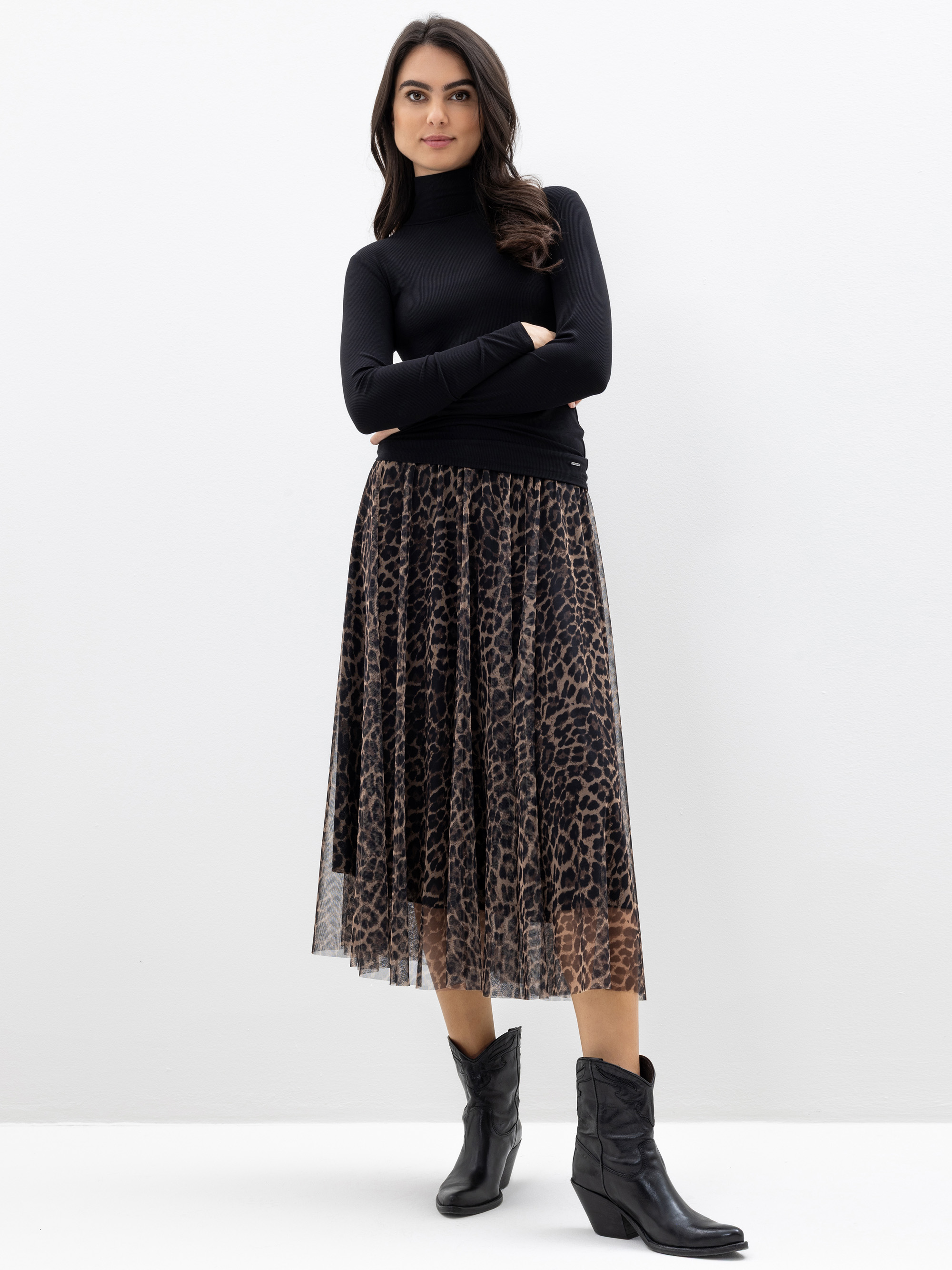 Eine Frau steht vor einer schlichten weißen Wand. Sie trägt einen schwarzen, langärmeligen Rollkragenpullover, einen mittellangen Rock mit Leopardenmuster und schwarze Ankle Boots. Sie hat langes dunkles Haar und die Arme verschränkt.