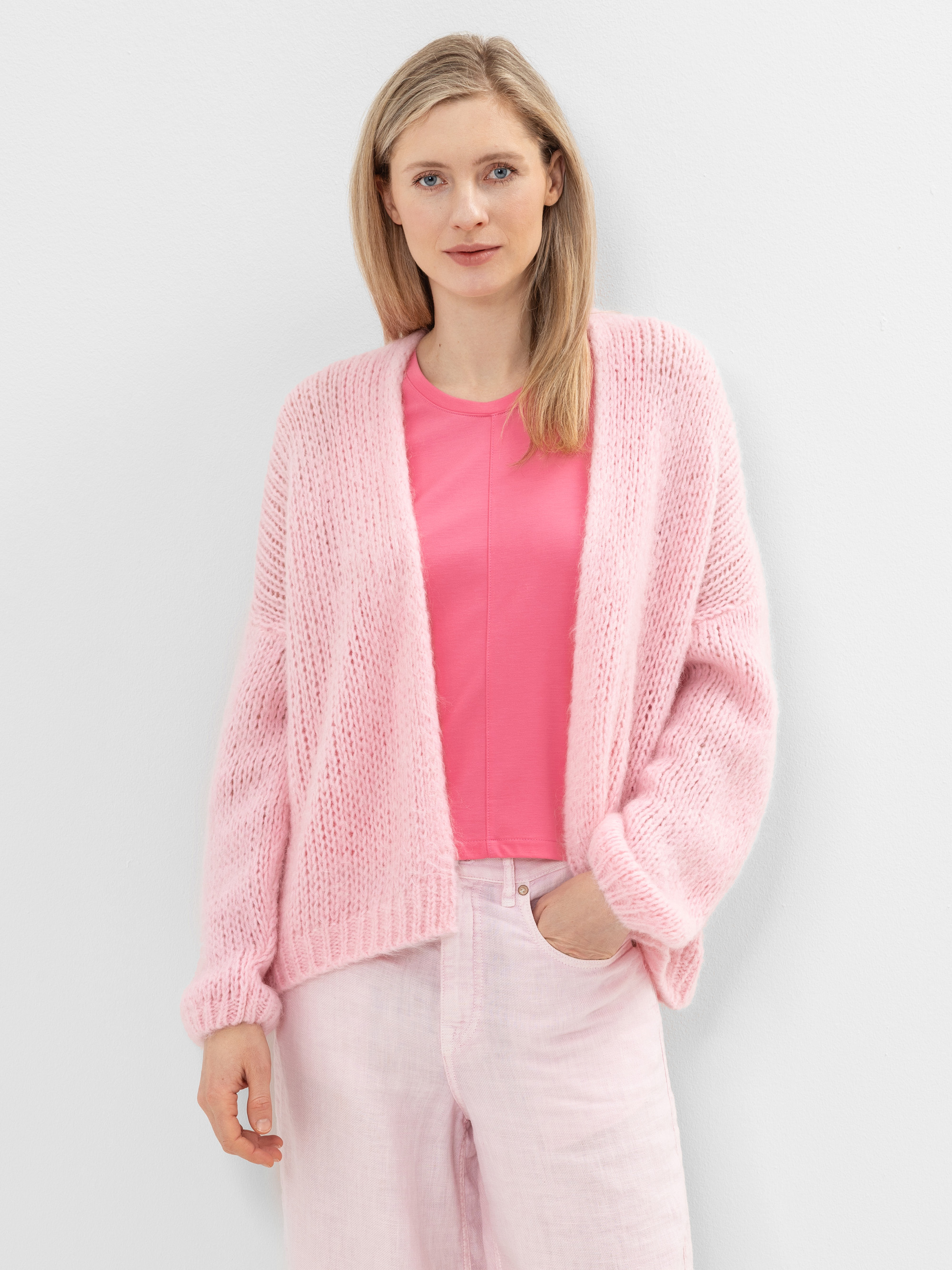 Eine Frau mit glattem blondem Haar steht vor einer weißen Wand und trägt eine hellrosa Strickjacke über einem rosa T-Shirt und eine hellrosa Hose. Sie hat eine Hand in der Tasche und einen entspannten Gesichtsausdruck.