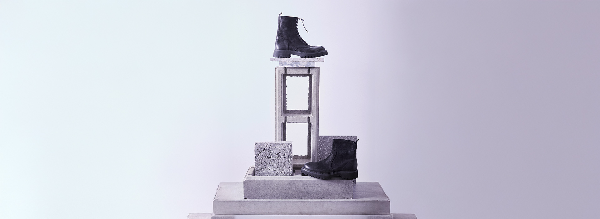 Ein Paar schwarzer Schnürstiefel auf gestapelten Betonblöcken und -platten, die in einer minimalistischen, geometrischen Komposition vor einem hellvioletten Hintergrund angeordnet sind.