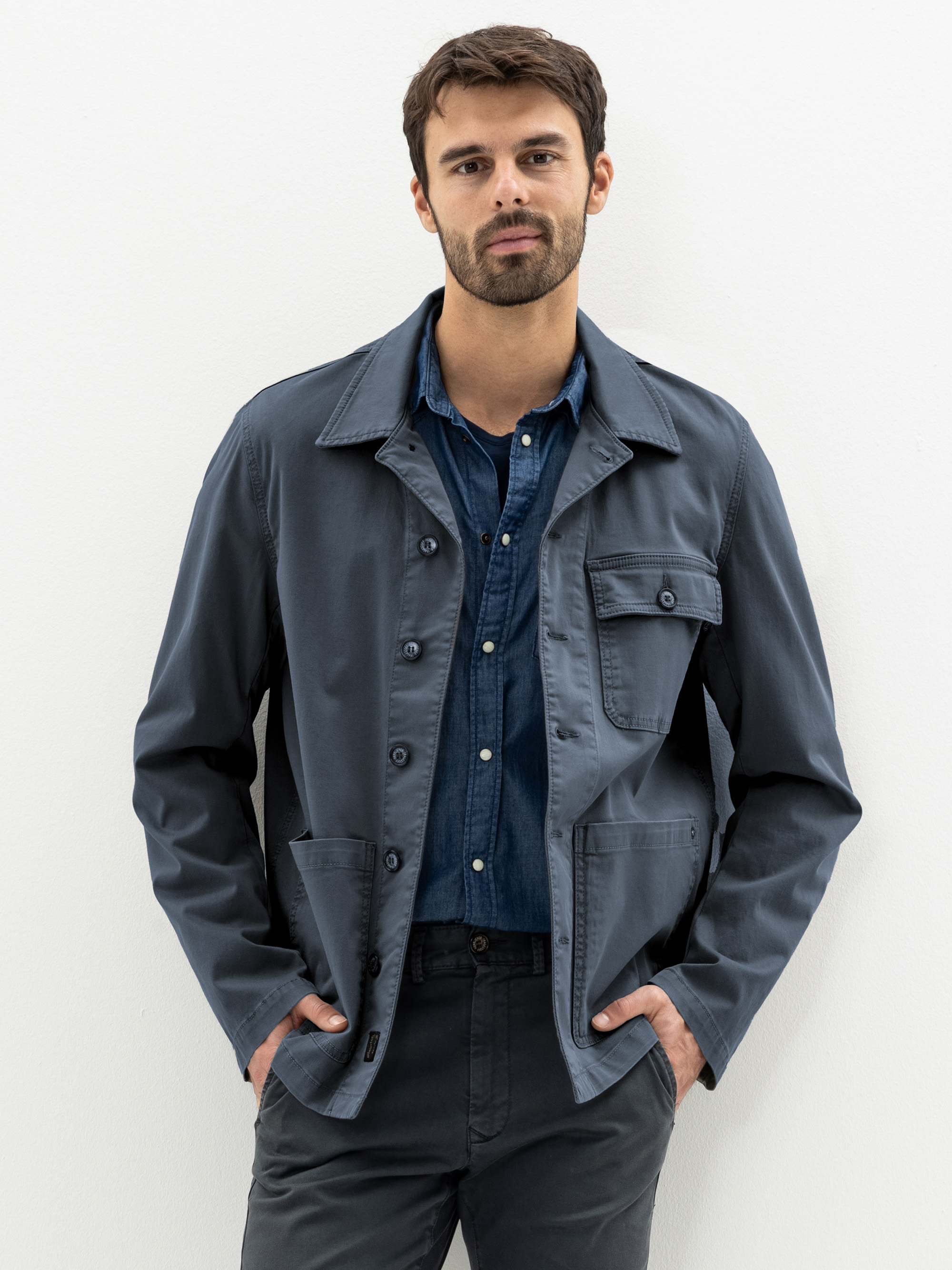 Ein Mann mit kurzen dunklen Haaren und einem Bart steht vor einem weißen Hintergrund. Er trägt ein blaues Button-up-Hemd unter einer grauen Jacke mit mehreren Taschen und eine dunkle Hose, die Hände in den Taschen.