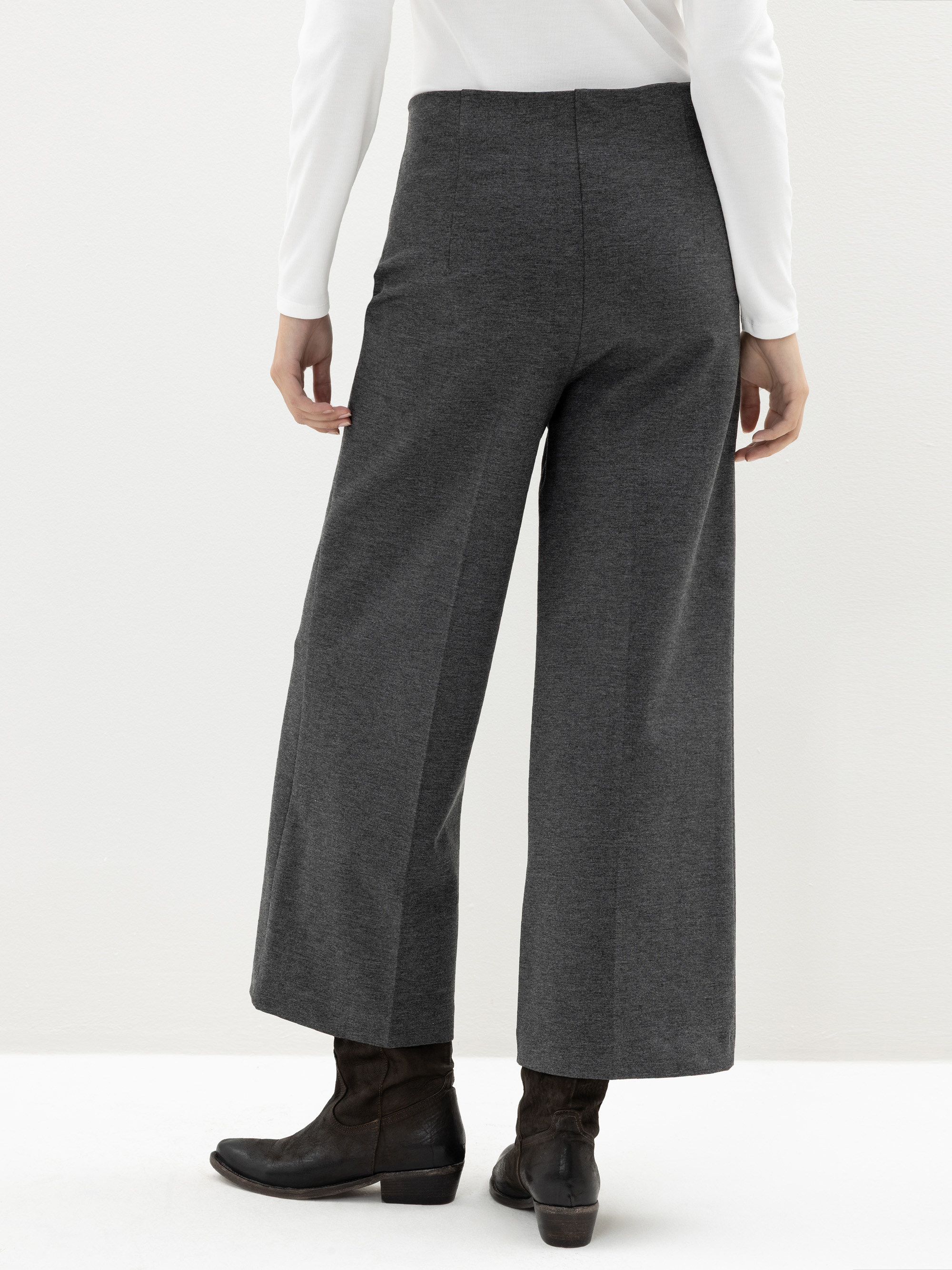 Eine Person steht mit dem Gesicht zur Wand, trägt ein weißes langärmeliges Oberteil, eine graue Hose mit weitem Bein und schwarze Stiefeletten vor einem schlichten hellen Hintergrund.
