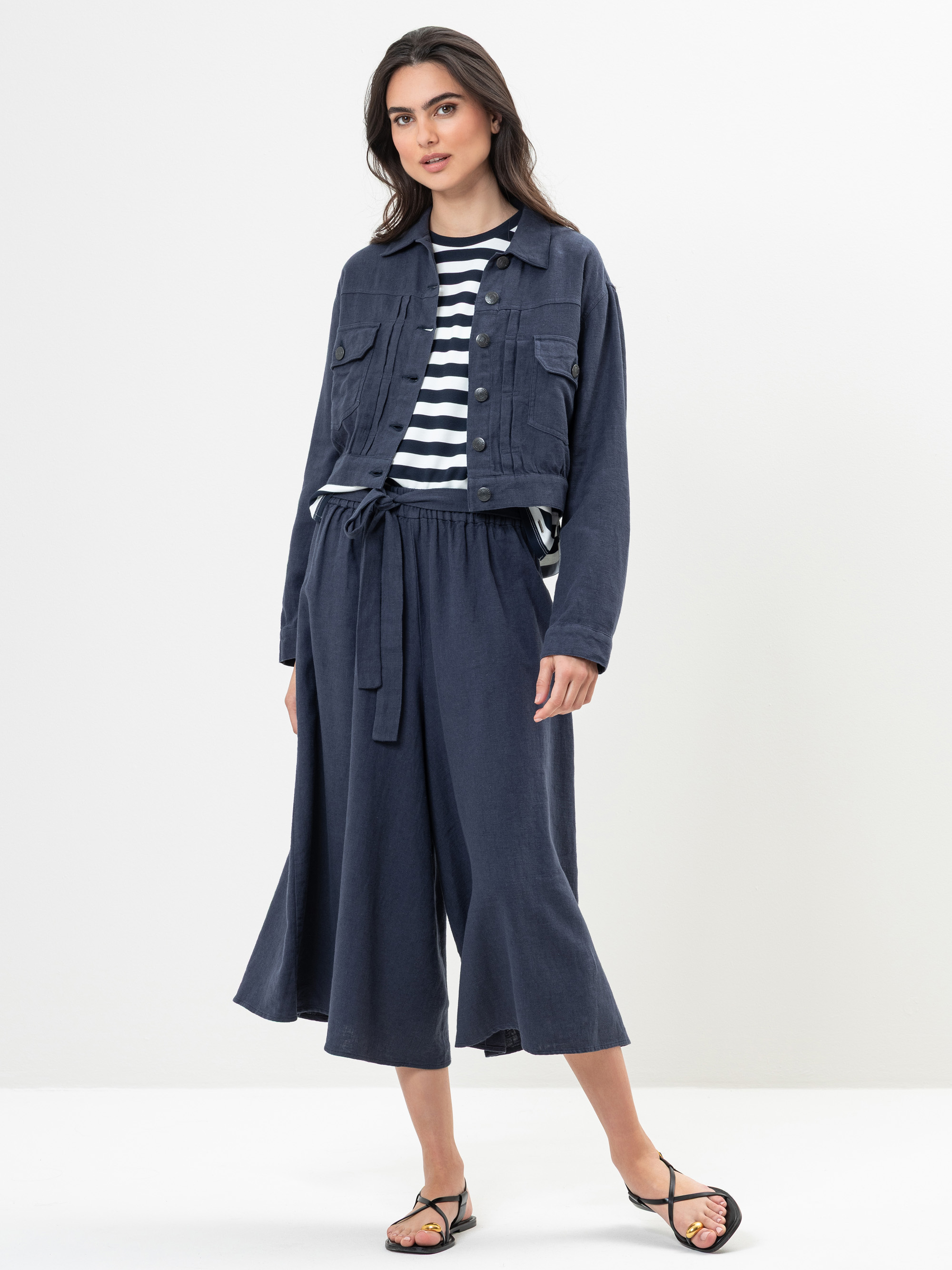 Eine Frau steht vor einem weißen Hintergrund und trägt eine marineblaue Jacke, ein schwarz-weiß gestreiftes Hemd, eine marineblaue Hose mit weitem Bein und Krawattenbund sowie schwarze Sandalen. Sie blickt mit entspanntem Gesichtsausdruck in die Kamera