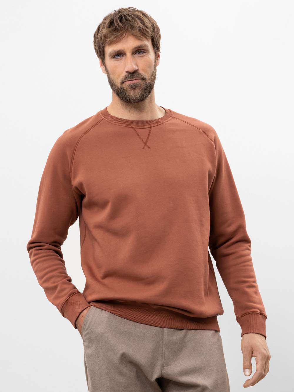 Ein Mann mit hellbraunem Haar und Bart, der ein rostfarbenes Sweatshirt und eine hellbraune Hose trägt, steht mit einer Hand in der Tasche vor einem schlichten weißen Hintergrund.