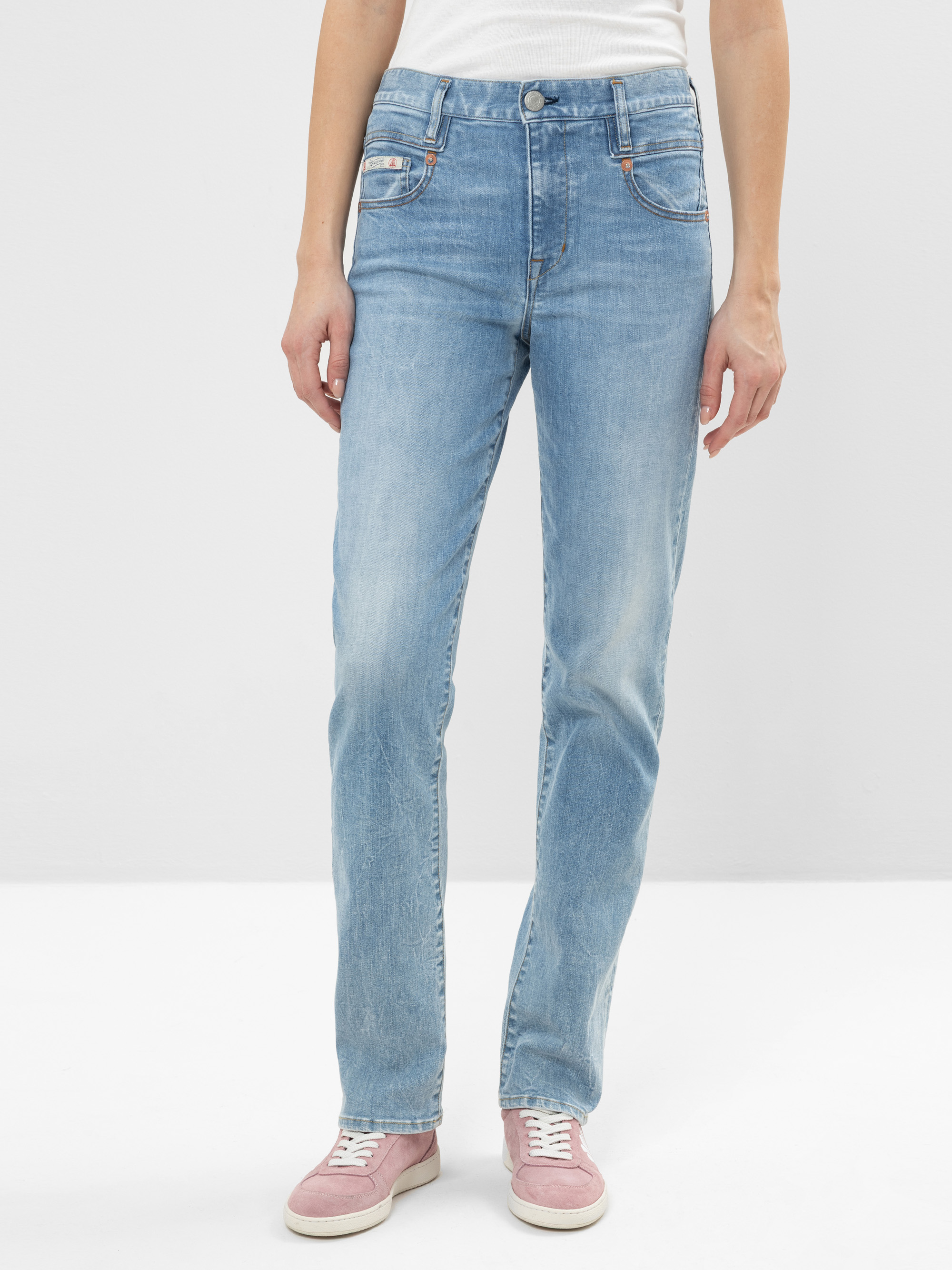 Eine Person, die hellblaue Jeans mit geradem Bein, ein weißes Hemd und rosa Turnschuhe trägt, steht vor einem schlichten weißen Hintergrund. Das Foto ist an den Schultern abgeschnitten und zeigt nur den unteren Teil des Oberkörpers und die Beine.