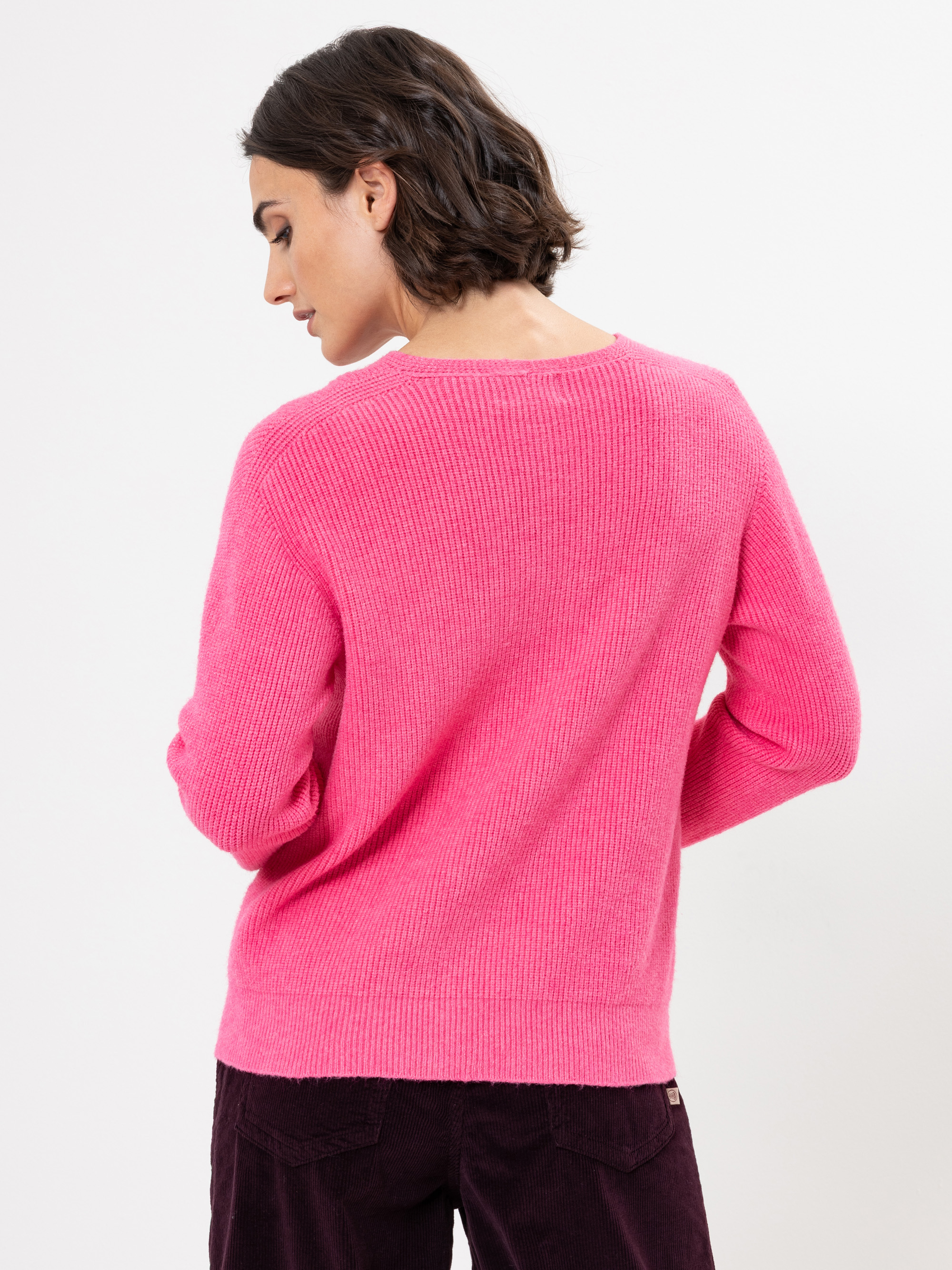 Eine Person mit kurzen braunen Haaren steht mit dem Rücken zur Kamera, trägt einen hellrosa, langärmeligen Strickpullover und eine dunkle Hose vor einem schlichten weißen Hintergrund.