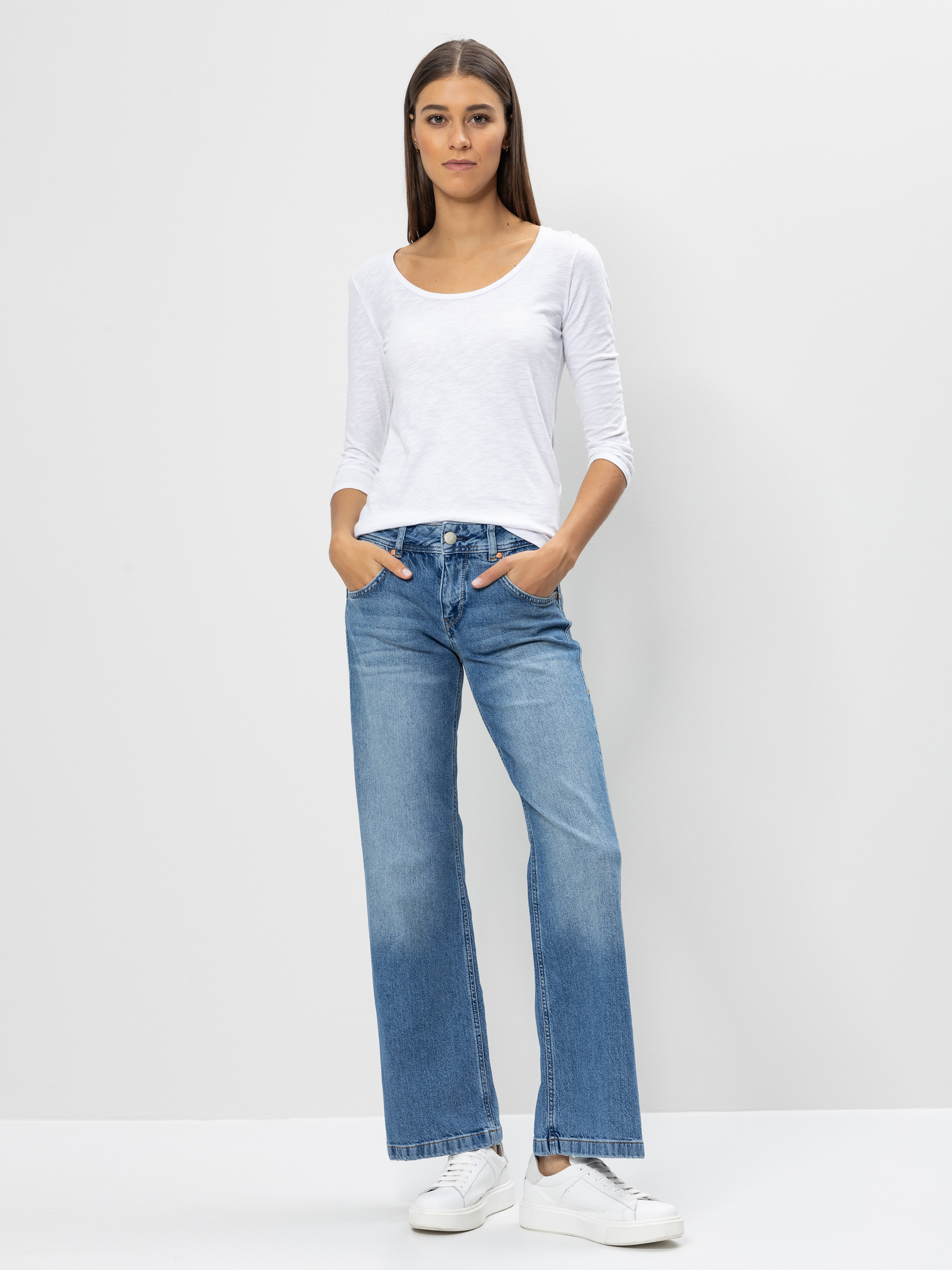 Eine Person mit langen braunen Haaren steht vor einem schlichten weißen Hintergrund, trägt ein weißes langärmeliges Oberteil, blaue Jeans mit hoher Taille und weiten Beinen und weiße Turnschuhe, die Hände in den Taschen und einen neutralen Gesichtsa