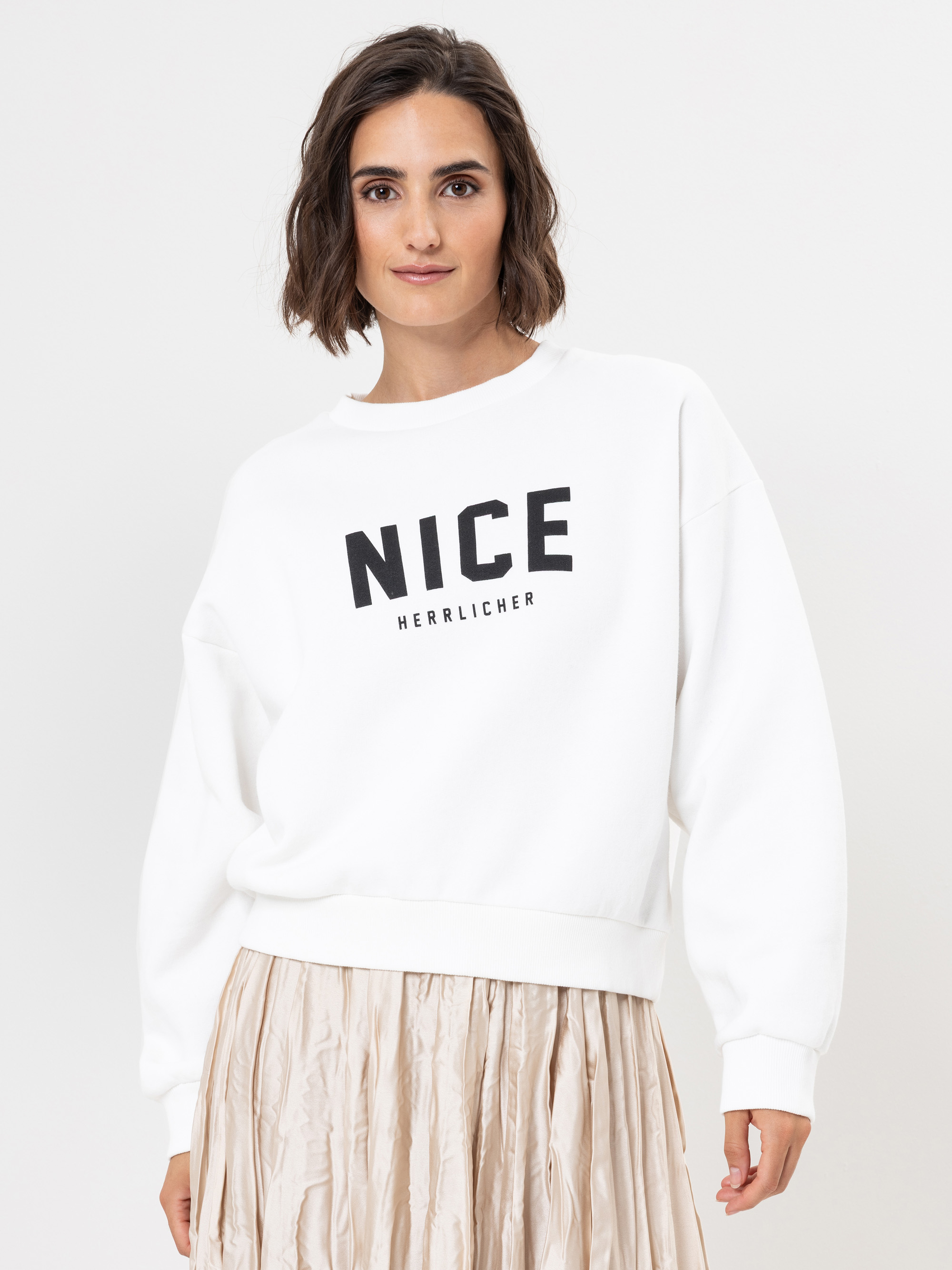 Eine Frau mit kurzen braunen Haaren trägt ein lockeres weißes Sweatshirt, auf dem in fetten schwarzen Buchstaben NICE HERRLICHER steht, gepaart mit einem beigen Faltenrock, und steht vor einem schlichten weißen Hintergrund.