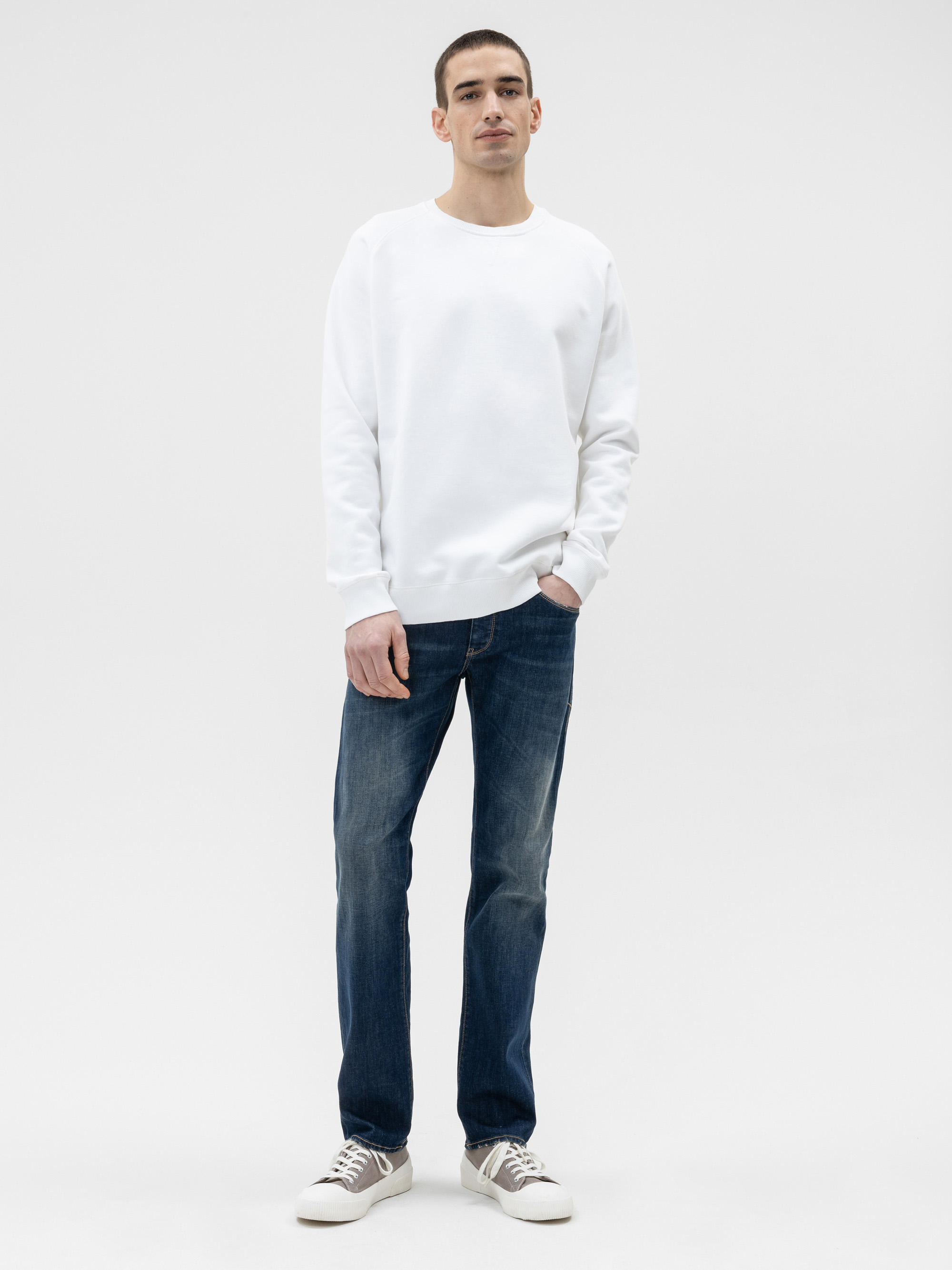 Ein Mann steht vor einem schlichten weißen Hintergrund, trägt ein weißes Sweatshirt, blaue Jeans und helle Turnschuhe, hat eine Hand in der Tasche und einen neutralen Gesichtsausdruck.