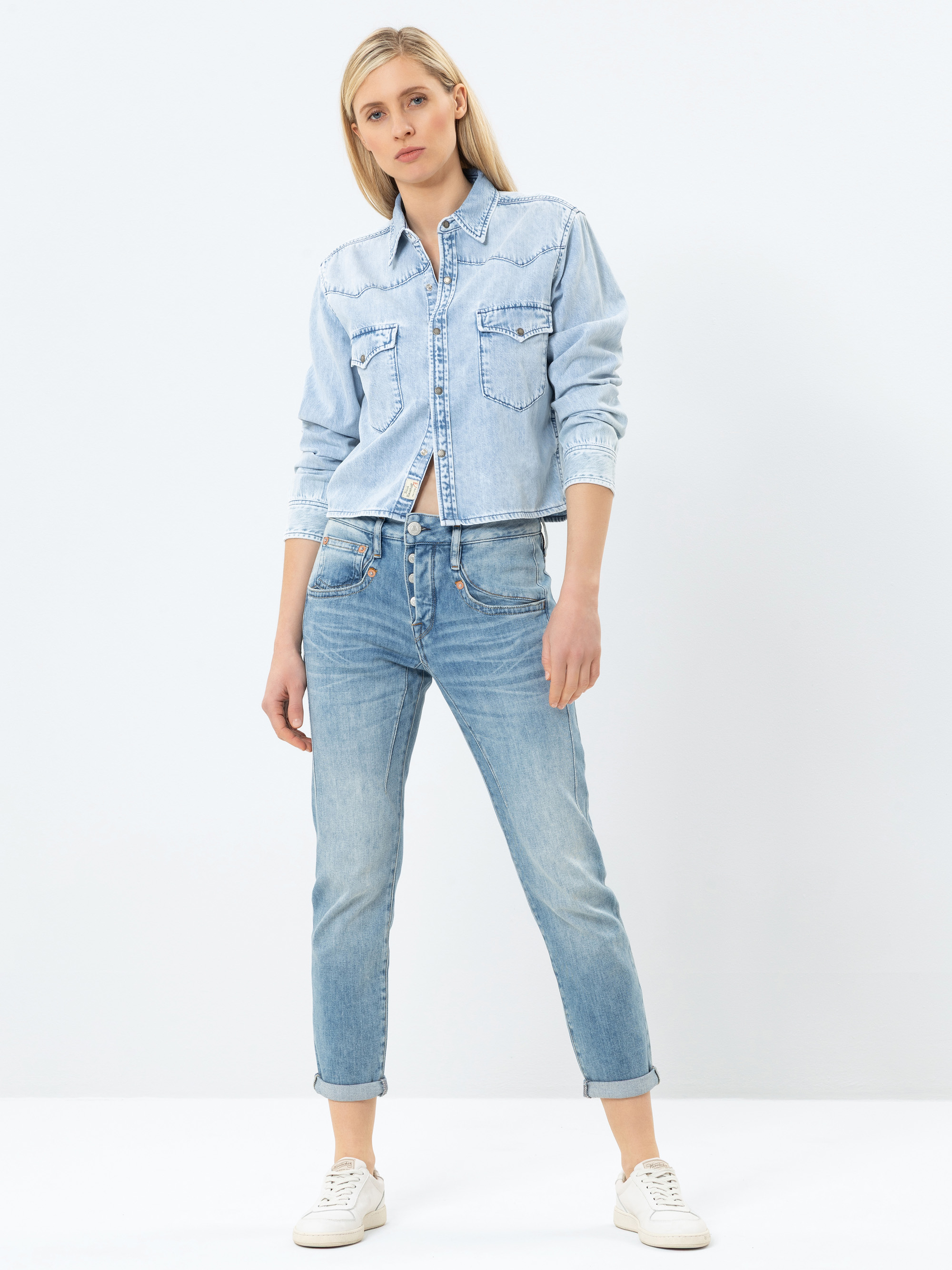 Eine Frau steht vor einem schlichten weißen Hintergrund und trägt ein hellblaues Jeanshemd, hellblaue Jeans mit Bündchen und weiße Turnschuhe. Sie hat glattes blondes Haar und eine entspannte, selbstbewusste Körperhaltung mit einem leicht angewinkelt