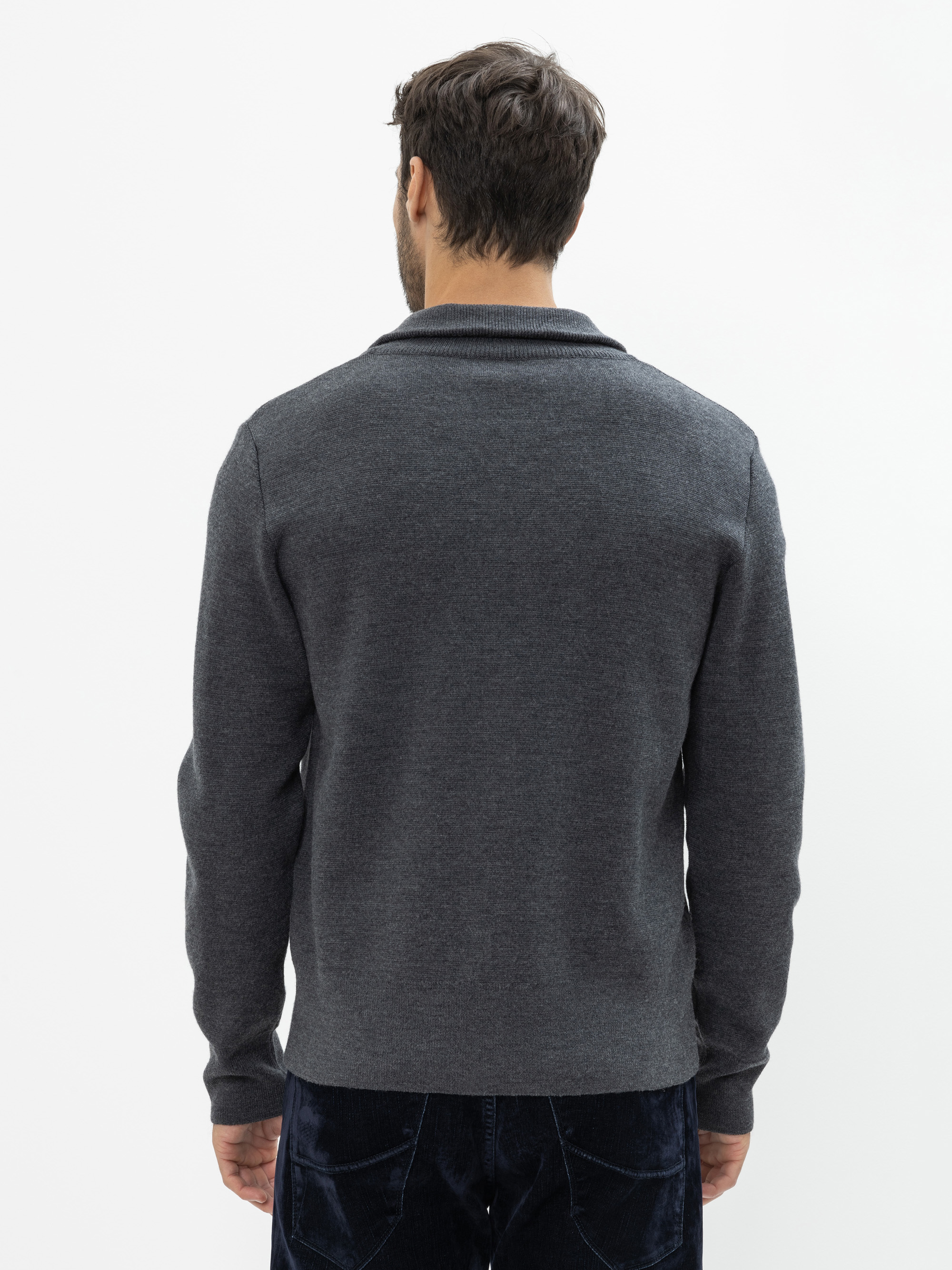 Ein Mann mit kurzen braunen Haaren steht mit dem Rücken zur Kamera, trägt einen dunkelgrauen langärmeligen Pullover und eine dunkle Hose vor einem schlichten weißen Hintergrund.