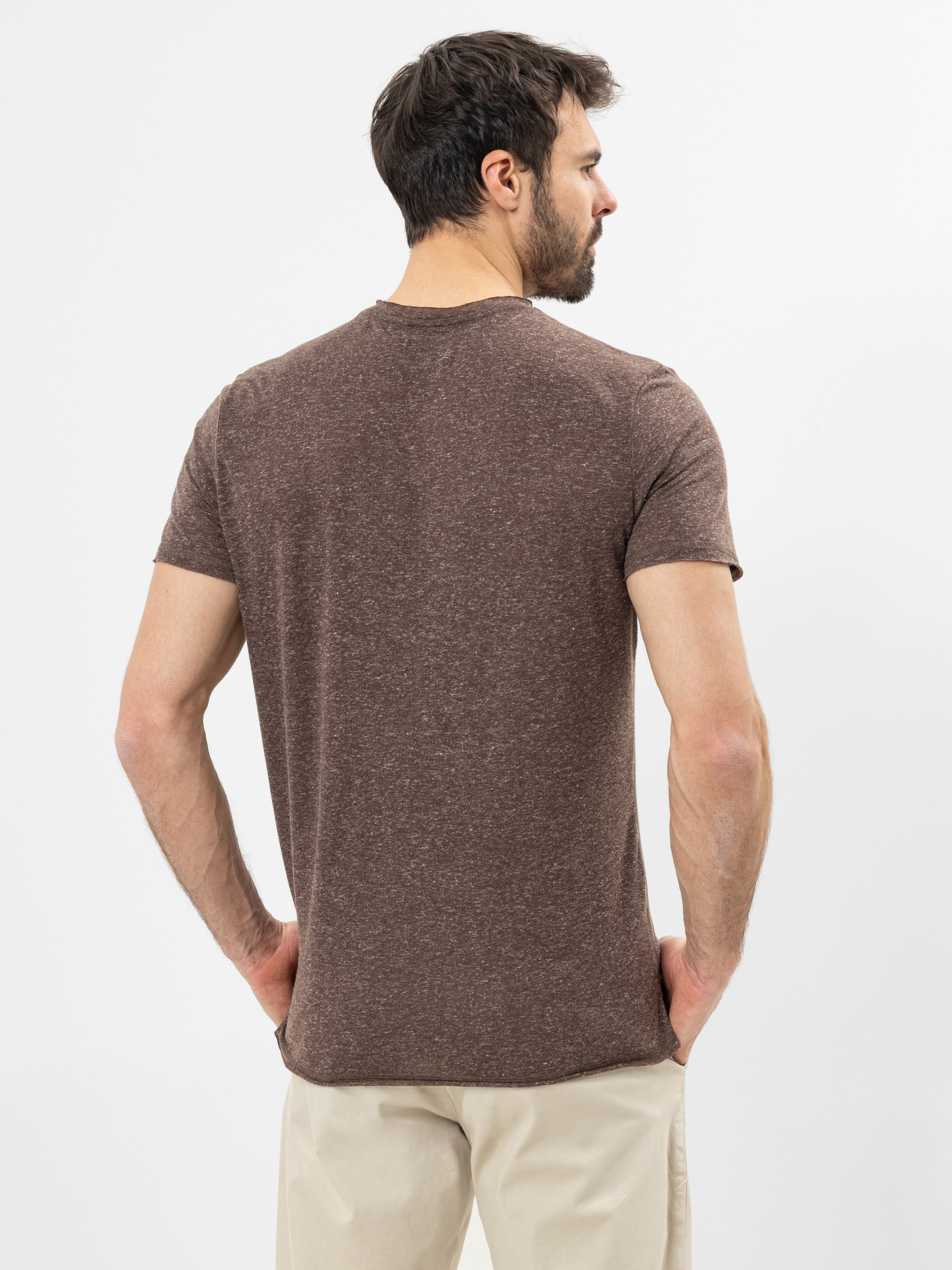 Ein Mann mit kurzen dunklen Haaren und einem Bart steht mit dem Rücken zur Kamera. Er trägt ein braunes kurzärmeliges T-Shirt und eine beigefarbene Hose vor einem schlichten weißen Hintergrund.