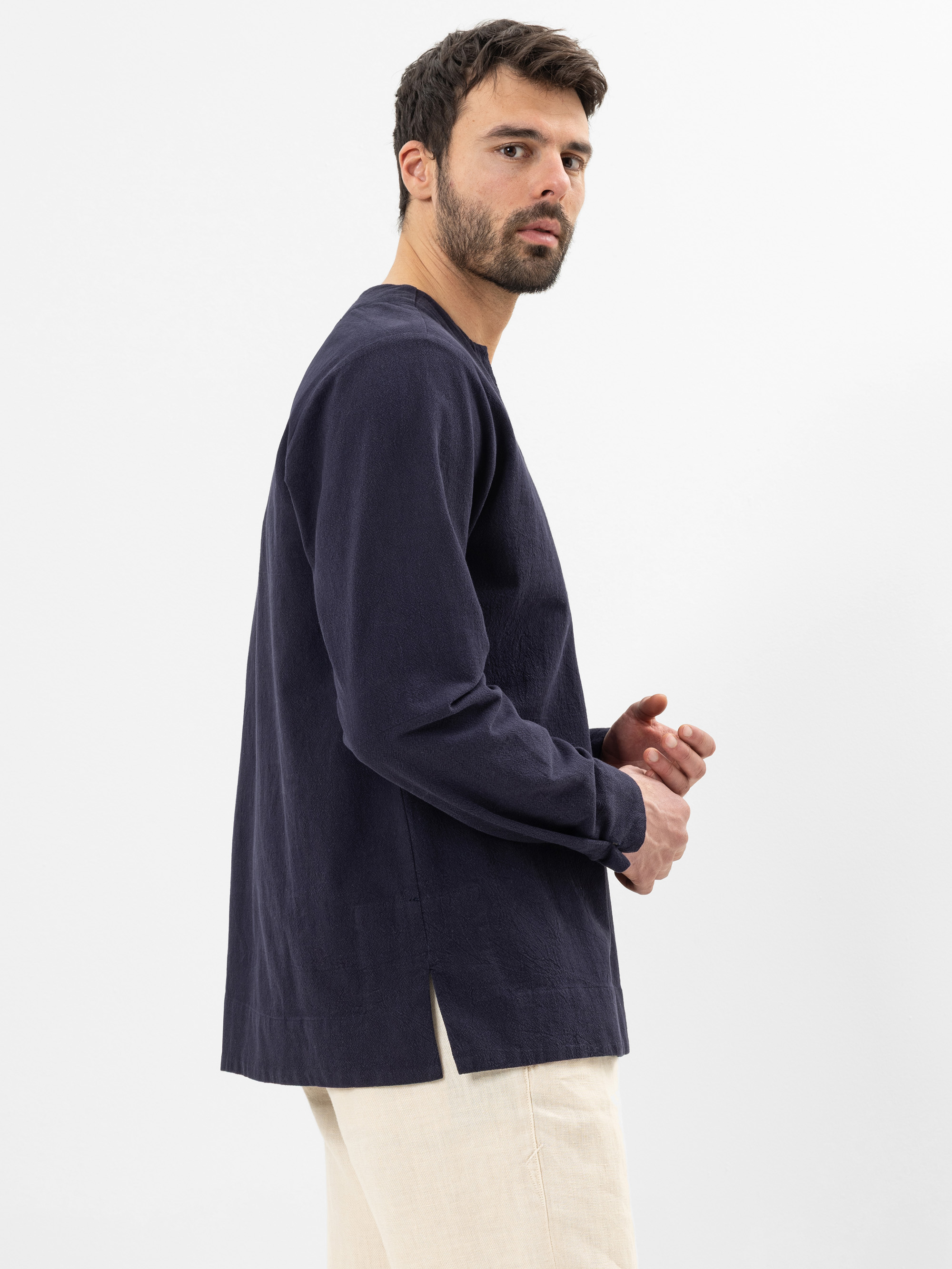 Ein Mann mit kurzen dunklen Haaren und einem Bart trägt ein locker sitzendes, langärmeliges marineblaues Hemd und eine hellbeige Hose. Er steht seitlich und schaut über seine Schulter vor einem schlichten weißen Hintergrund.