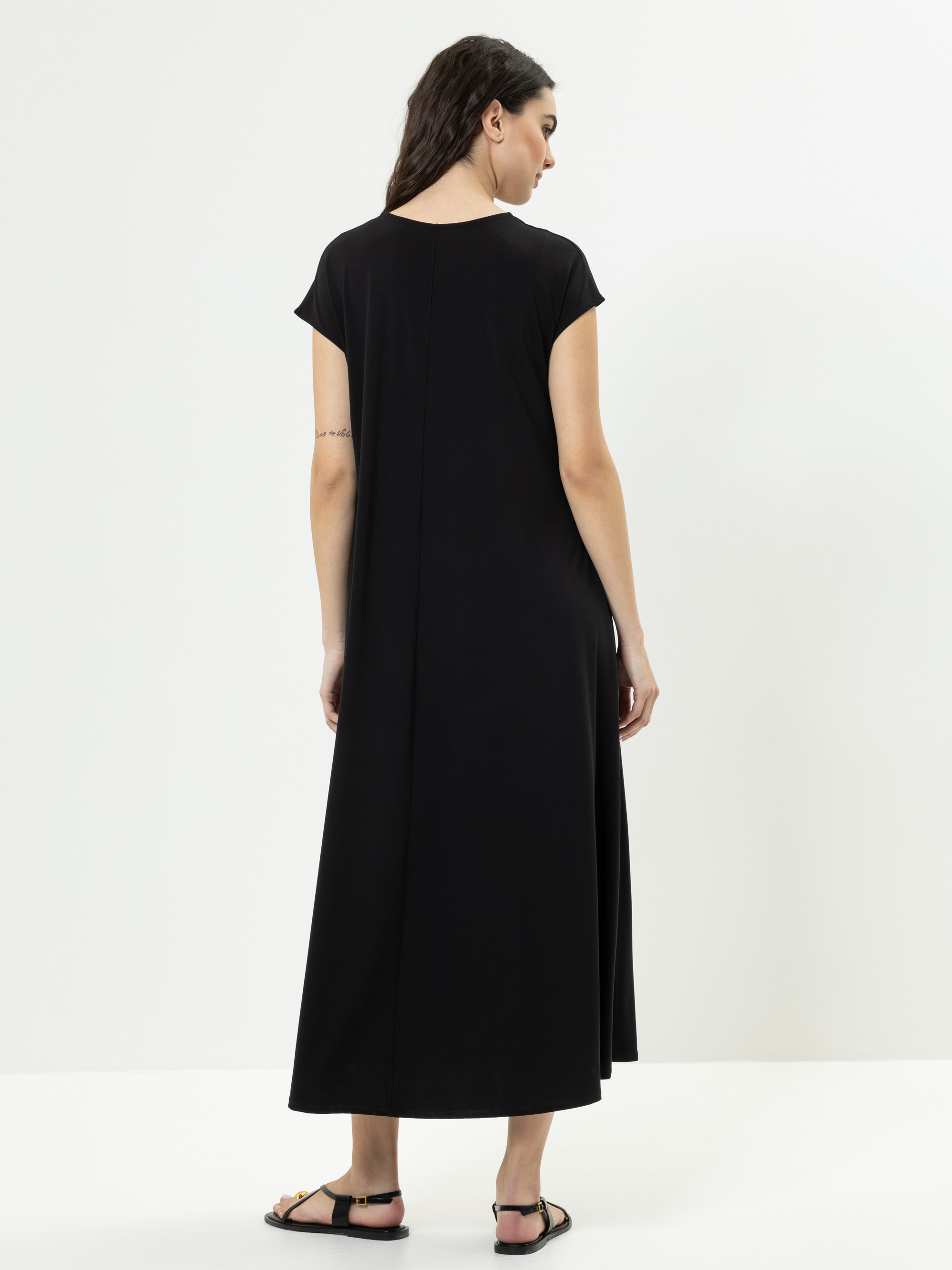 Eine Frau steht mit dem Gesicht nach hinten, trägt ein schwarzes, kurzärmeliges, knöchellanges Kleid und schwarze Sandalen. Ihr Haar ist offen und sie hat eine Tätowierung auf ihrem linken Oberarm. Der Hintergrund ist einfarbig weiß.