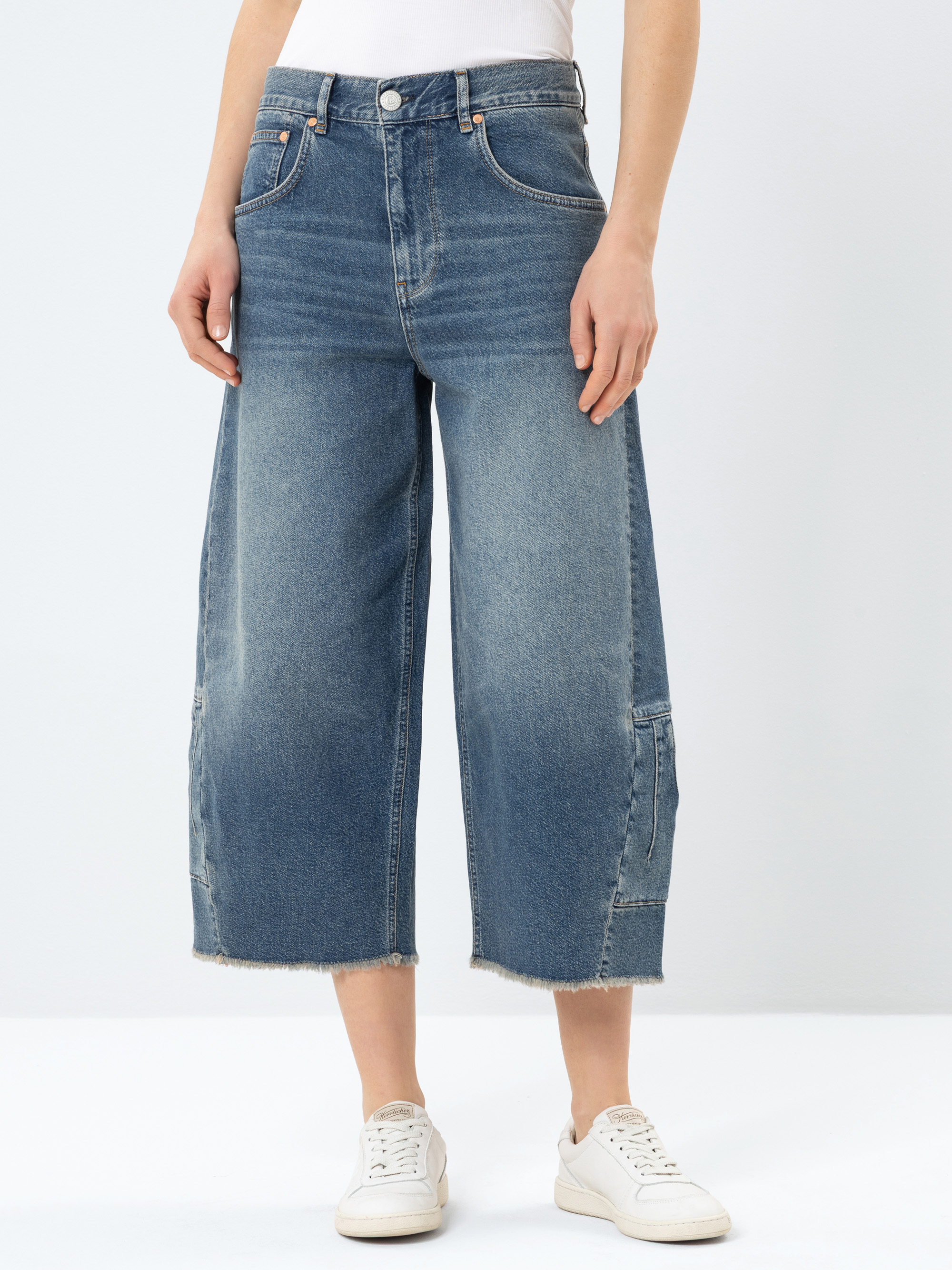 Eine Person, die weit geschnittene blaue Jeans mit ausgefransten Säumen, ein weißes Oberteil und weiße Turnschuhe trägt, steht vor einem schlichten hellen Hintergrund. Das Foto ist an den Schultern abgeschnitten.