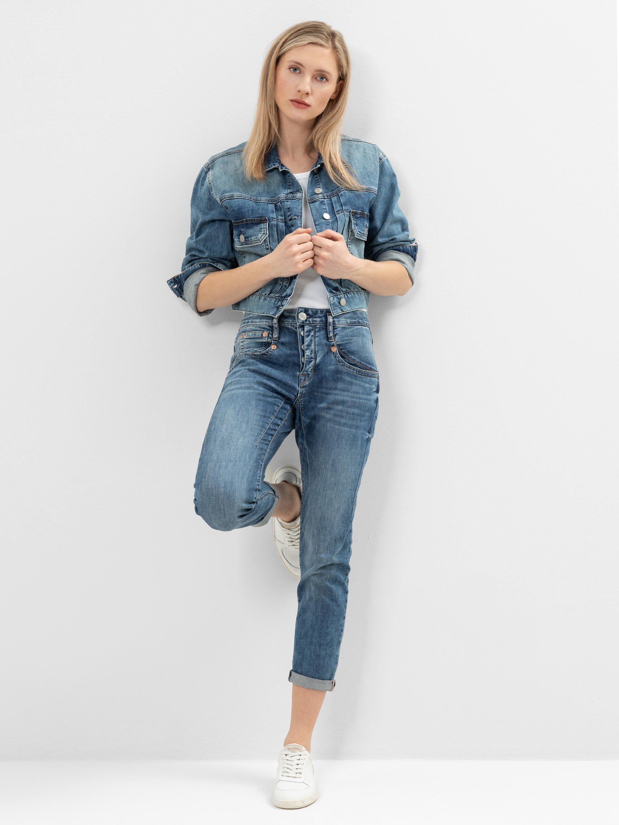 Eine Frau, die eine blaue Jeansjacke und Jeans trägt, steht mit einem angewinkelten Bein an einer weißen Wand und stützt sich mit dem Fuß auf der Wand ab. Sie hat helles Haar und trägt weiße Turnschuhe.