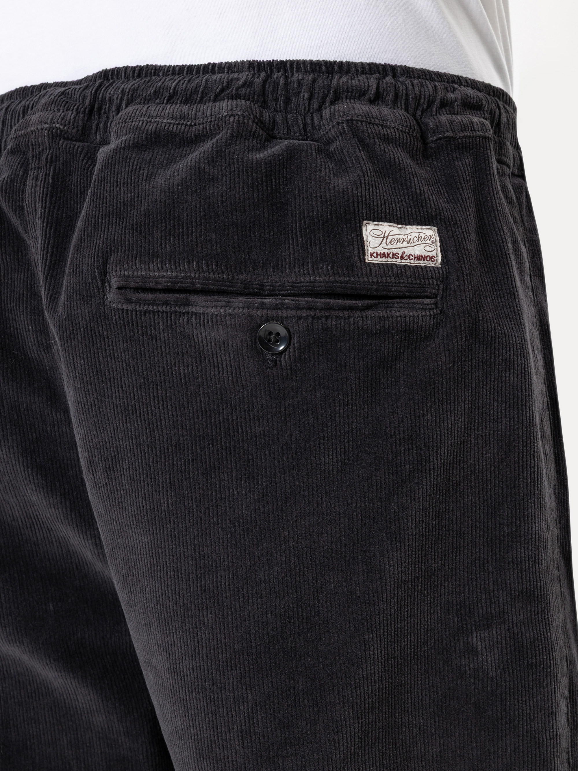 Nahaufnahme einer Person, die eine dunkle Kordhose mit elastischem Bund und einer Gesäßtasche mit Knopf trägt. Über der Tasche ist ein kleines weißes Aufnäher-Etikett aufgenäht, und oben ist ein weißes Hemd teilweise sichtbar.