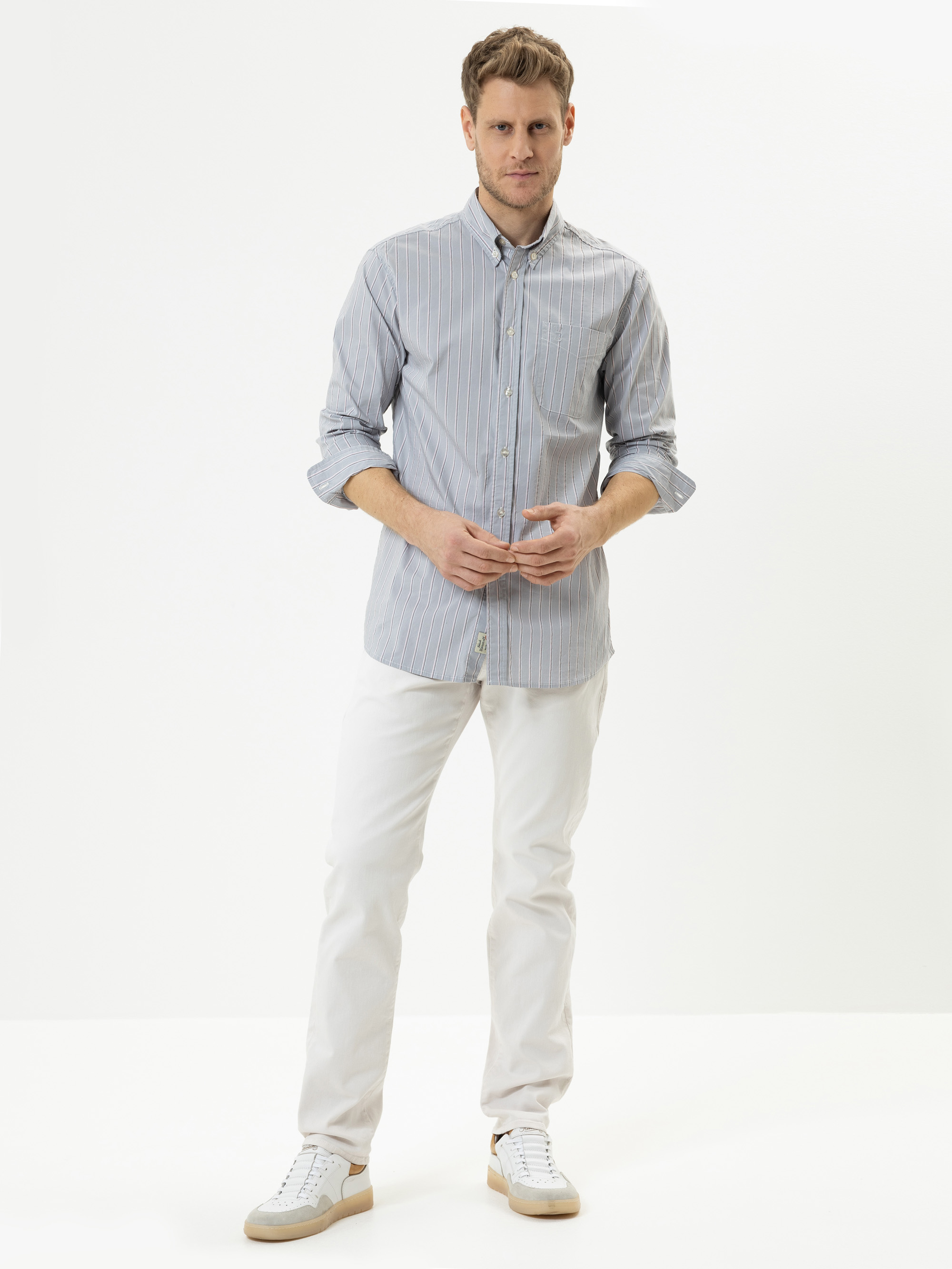 Ein Mann steht vor einem schlichten weißen Hintergrund, trägt ein hellblau gestreiftes Button-up-Hemd, eine weiße Hose und weiße Turnschuhe und schaut mit neutralem Blick direkt in die Kamera.