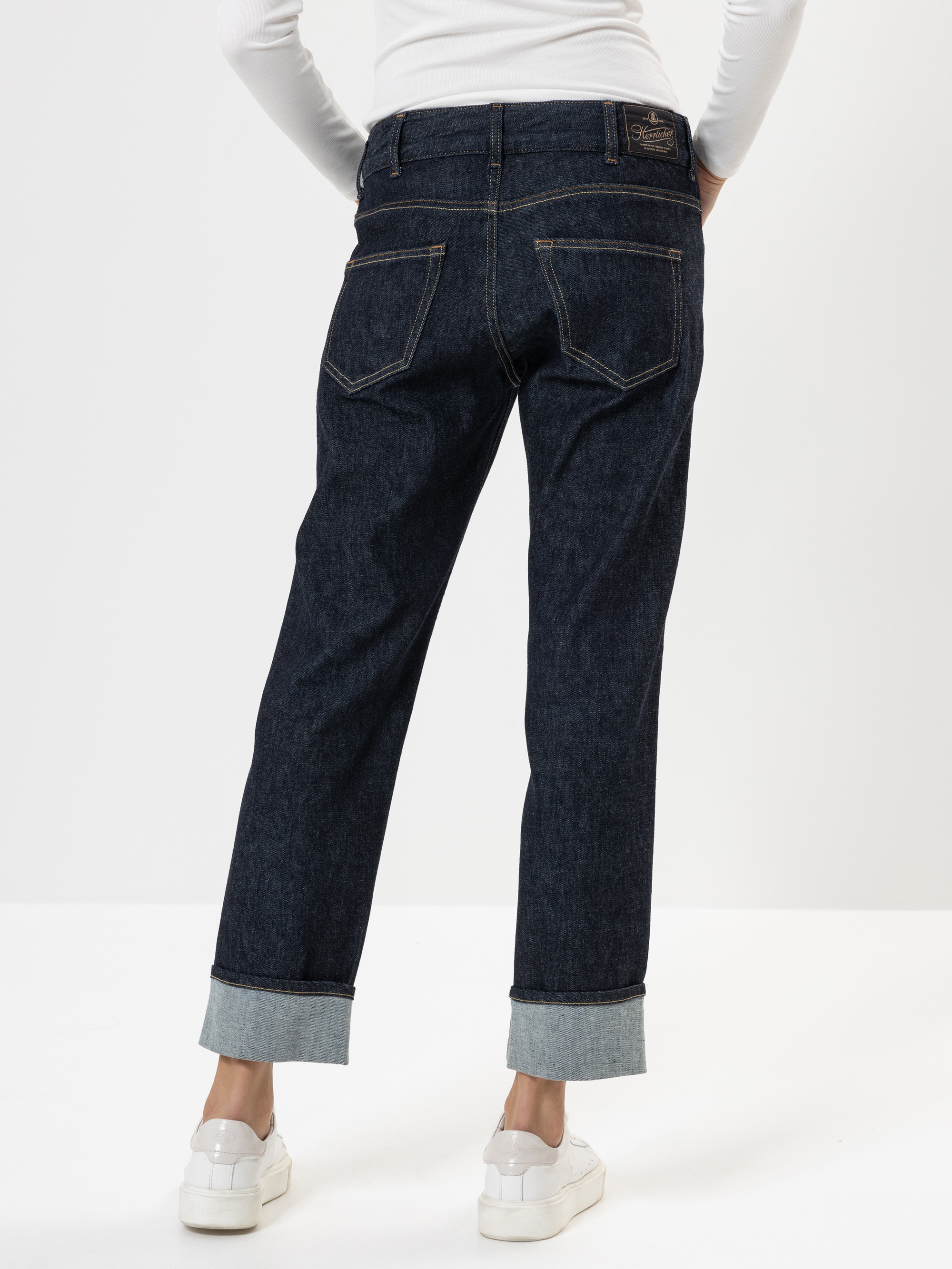 Eine Person, die dunkelblaue Jeans mit Bündchen, ein weißes langärmeliges Oberteil und weiße Turnschuhe trägt, steht mit dem Rücken zur Kamera vor einem schlichten weißen Hintergrund.