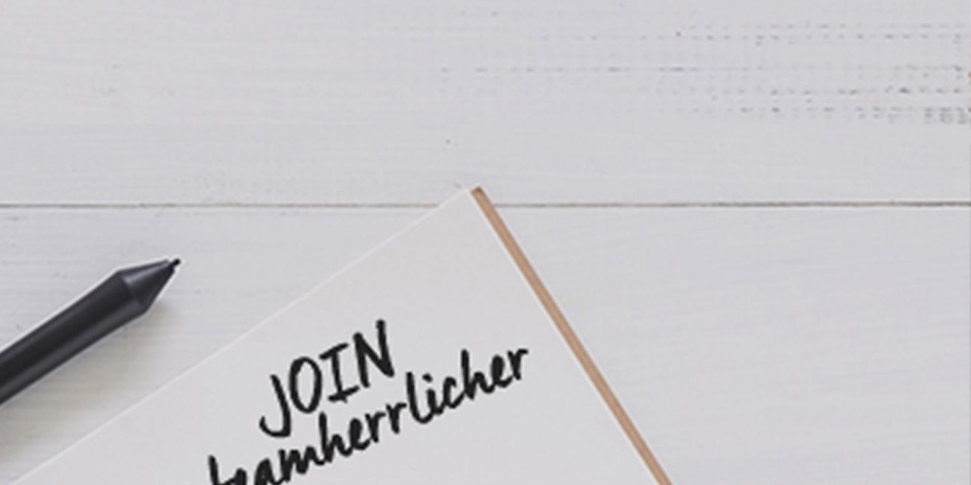 Ein schwarzer Stift liegt neben einem aufgeschlagenen Notizbuch auf einer weißen Holzfläche. Auf der sichtbaren Seite des Notizbuchs steht in großen Buchstaben JOIN, gefolgt von einem teilweise sichtbaren, handgeschriebenen Text.