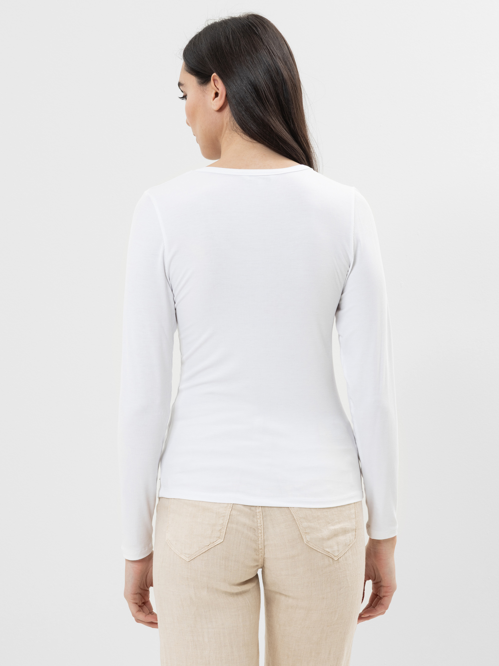 Eine Frau mit langen dunklen Haaren, von hinten gesehen, trägt ein weißes, langärmeliges Hemd und eine hellbeige Hose und steht vor einem schlichten weißen Hintergrund.
