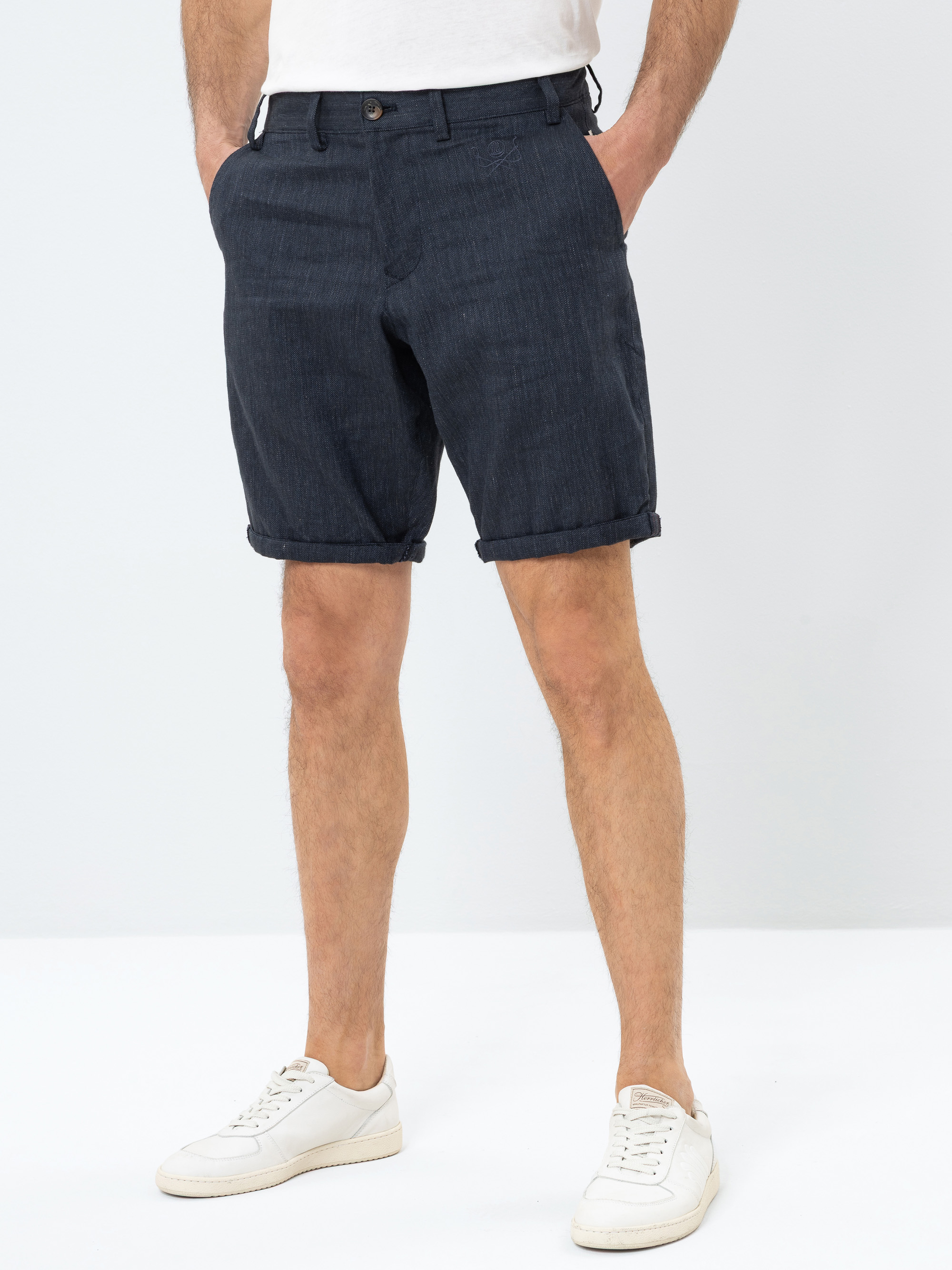Ein Mann steht mit den Händen in den Taschen, trägt dunkelblaue, gekrempelte Shorts und weiße Turnschuhe vor einem schlichten weißen Hintergrund. Nur der untere Teil seines Oberkörpers und seine Beine sind zu sehen.