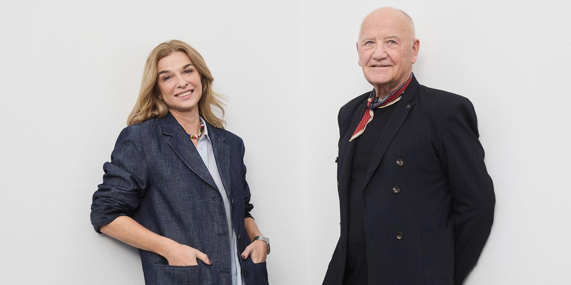 Eine Frau und ein älterer Mann stehen vor einem schlichten weißen Hintergrund, beide lächeln. Die Frau trägt einen blauen Blazer und hat die Hände in den Taschen. Der Mann trägt einen dunklen Anzug mit einem roten Schal und hat die Hände hinter dem