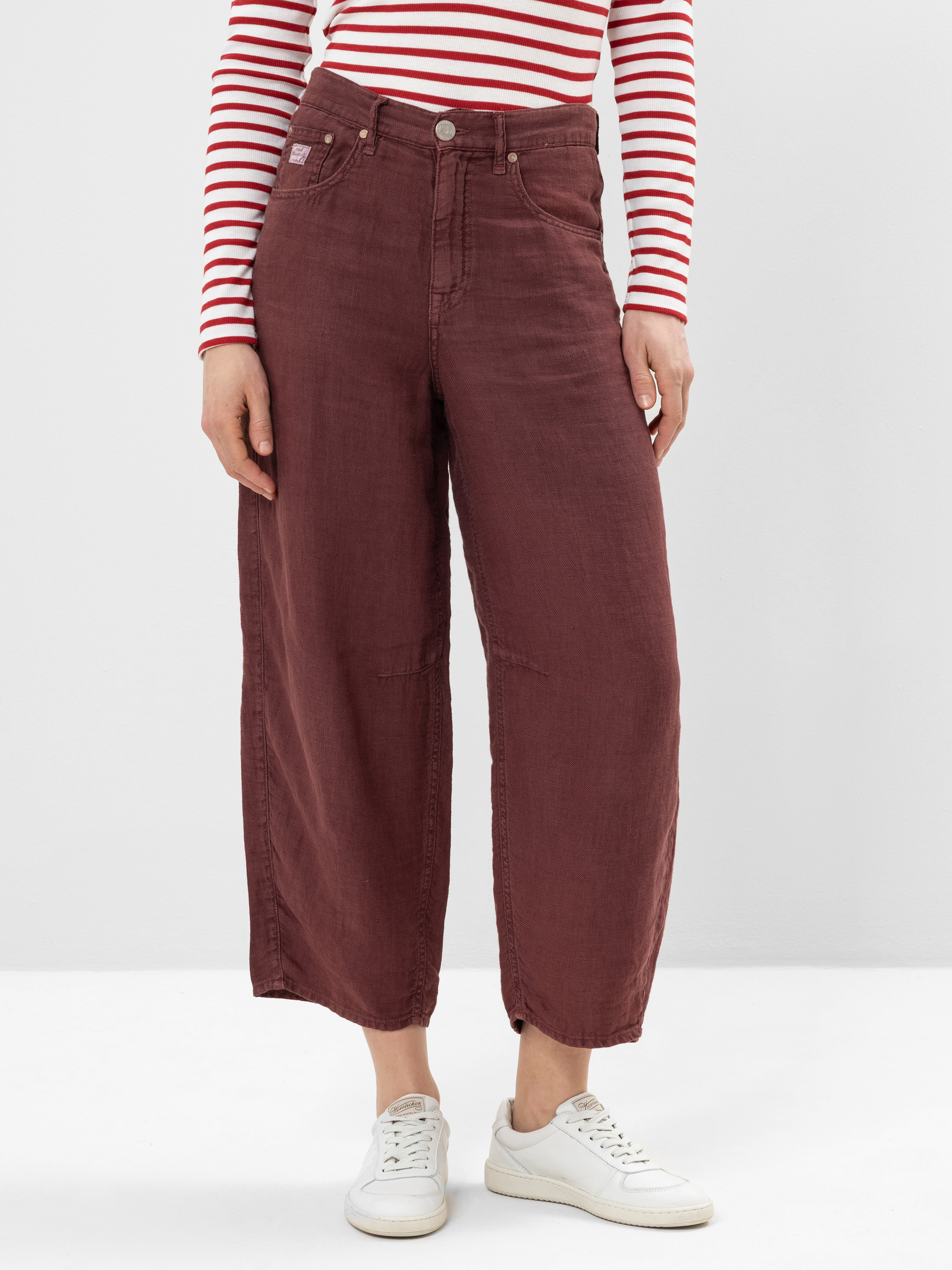 Eine Person, die eine kastanienbraune Hose mit weiten Beinen, ein langärmeliges rot-weiß gestreiftes Hemd und weiße Turnschuhe trägt, steht vor einem schlichten weißen Hintergrund.