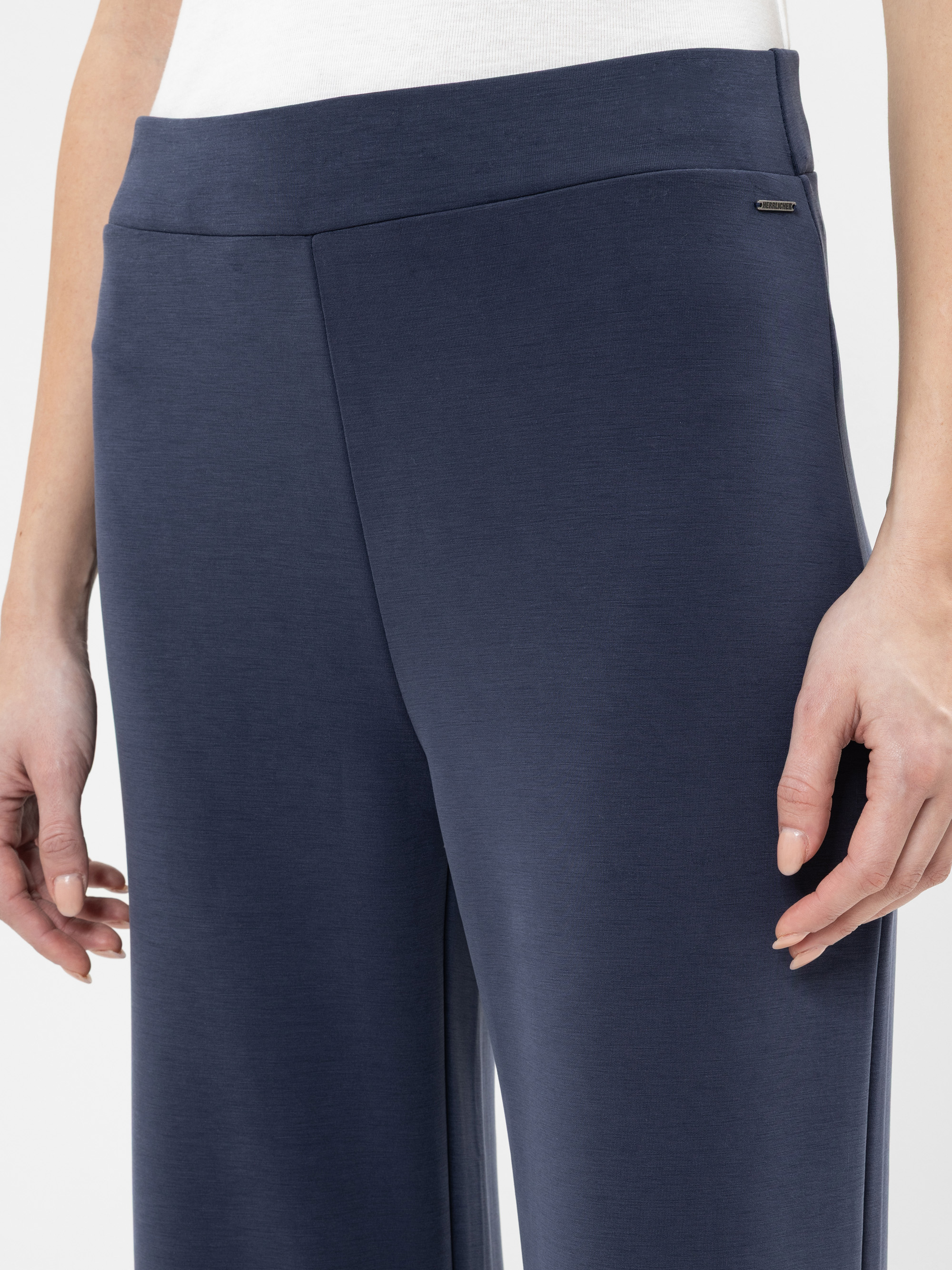 Eine Person, die eine marineblaue Hose mit weitem Bein und ein weißes Oberteil trägt, wobei die linke Hand an der Seite ruht, wird von der Taille bis zur Mitte des Oberschenkels vor einem einfarbigen Hintergrund gezeigt.