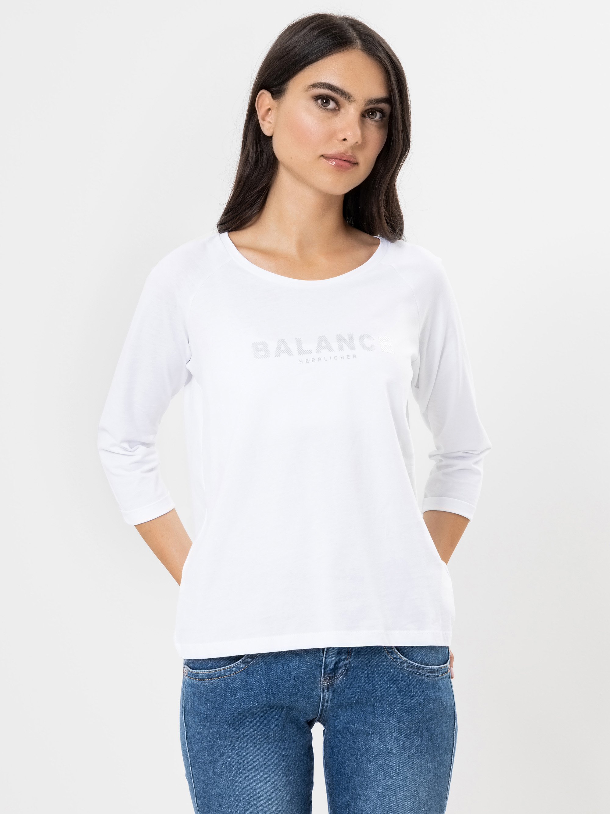 Eine Frau mit langen dunklen Haaren steht vor einem schlichten weißen Hintergrund, trägt ein weißes langärmeliges Hemd mit der Aufschrift BALANCE und blaue Jeans, die Hände auf dem Rücken.